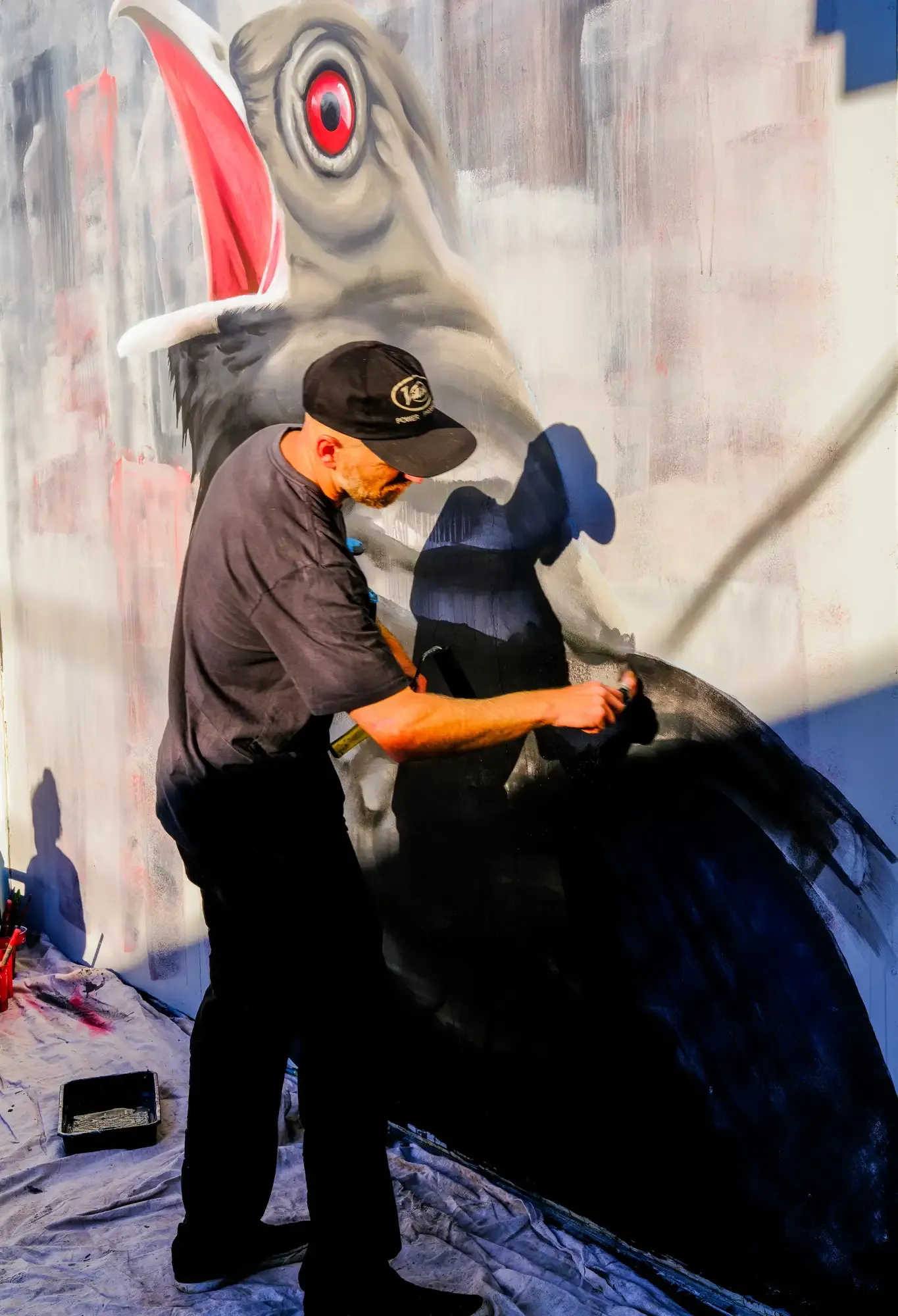 Artist painting a large bird mural on a wall, wearing a black cap and shirt, with sunlight casting shadows.