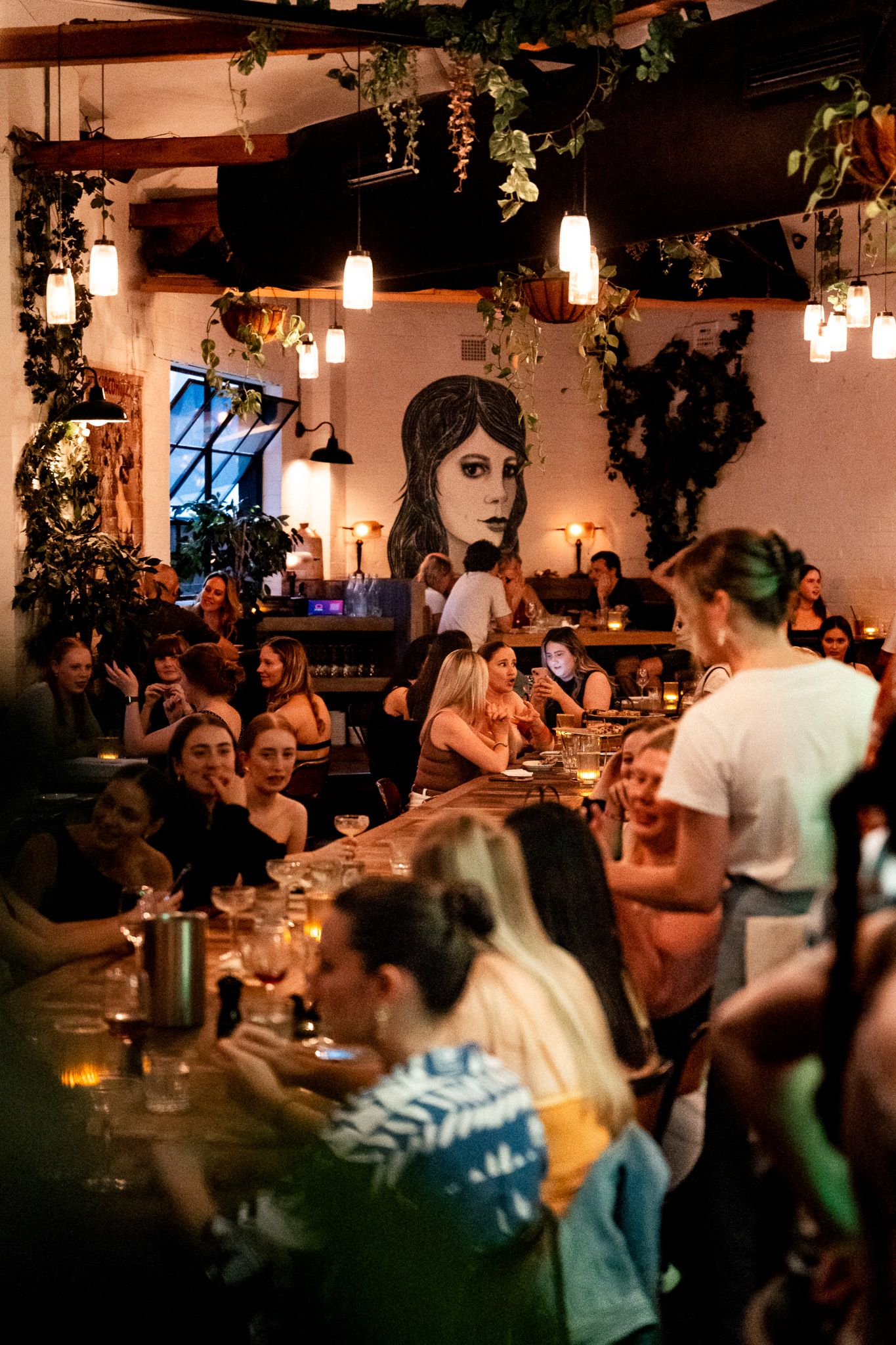 A lively, crowded restaurant with people dining and socializing. Warm lighting and wall art create a cozy atmosphere. Hanging plants add greenery.