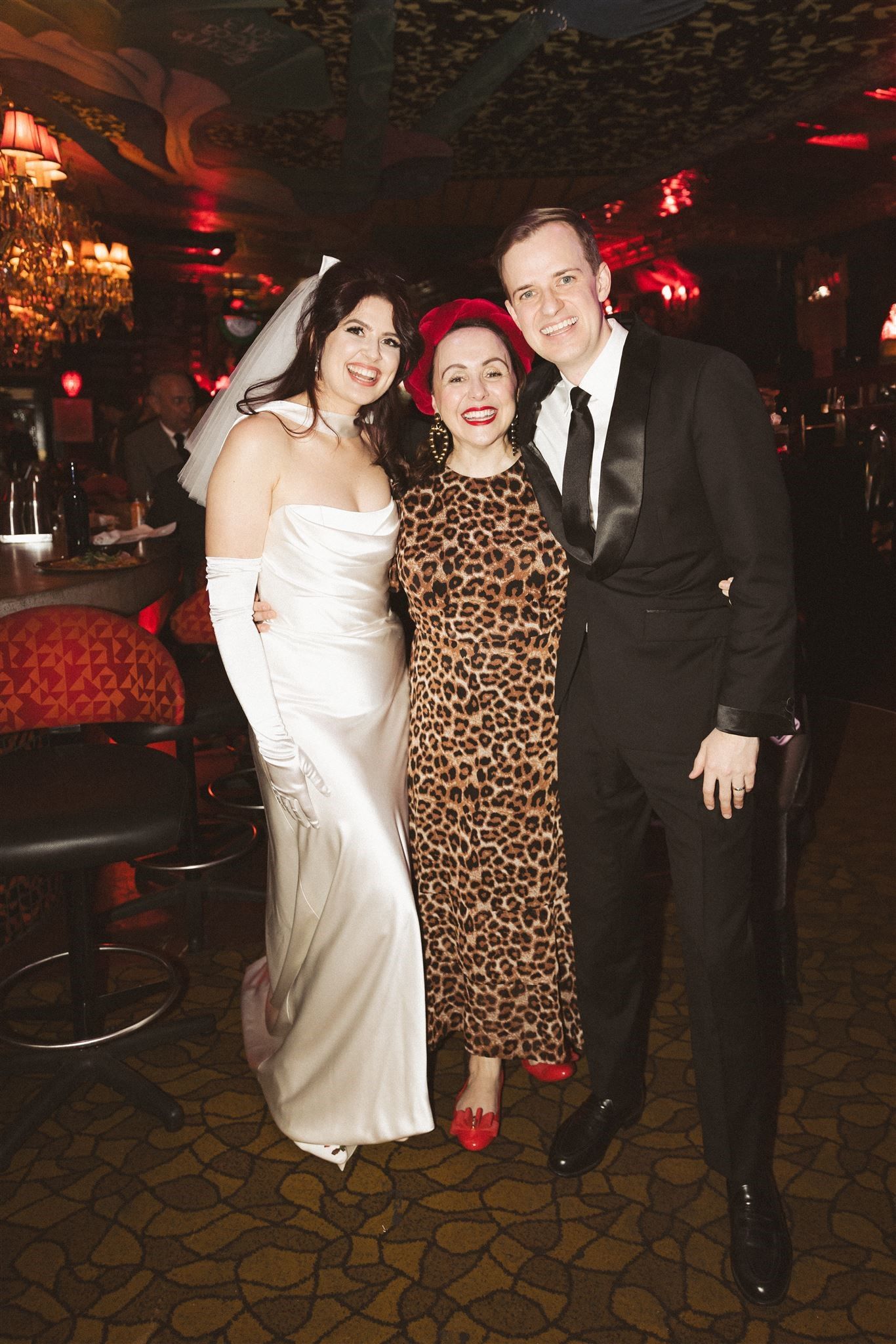 A bride in a white gown, a person in a leopard print dress, and a man in a tuxedo smile together in a warmly lit venue.