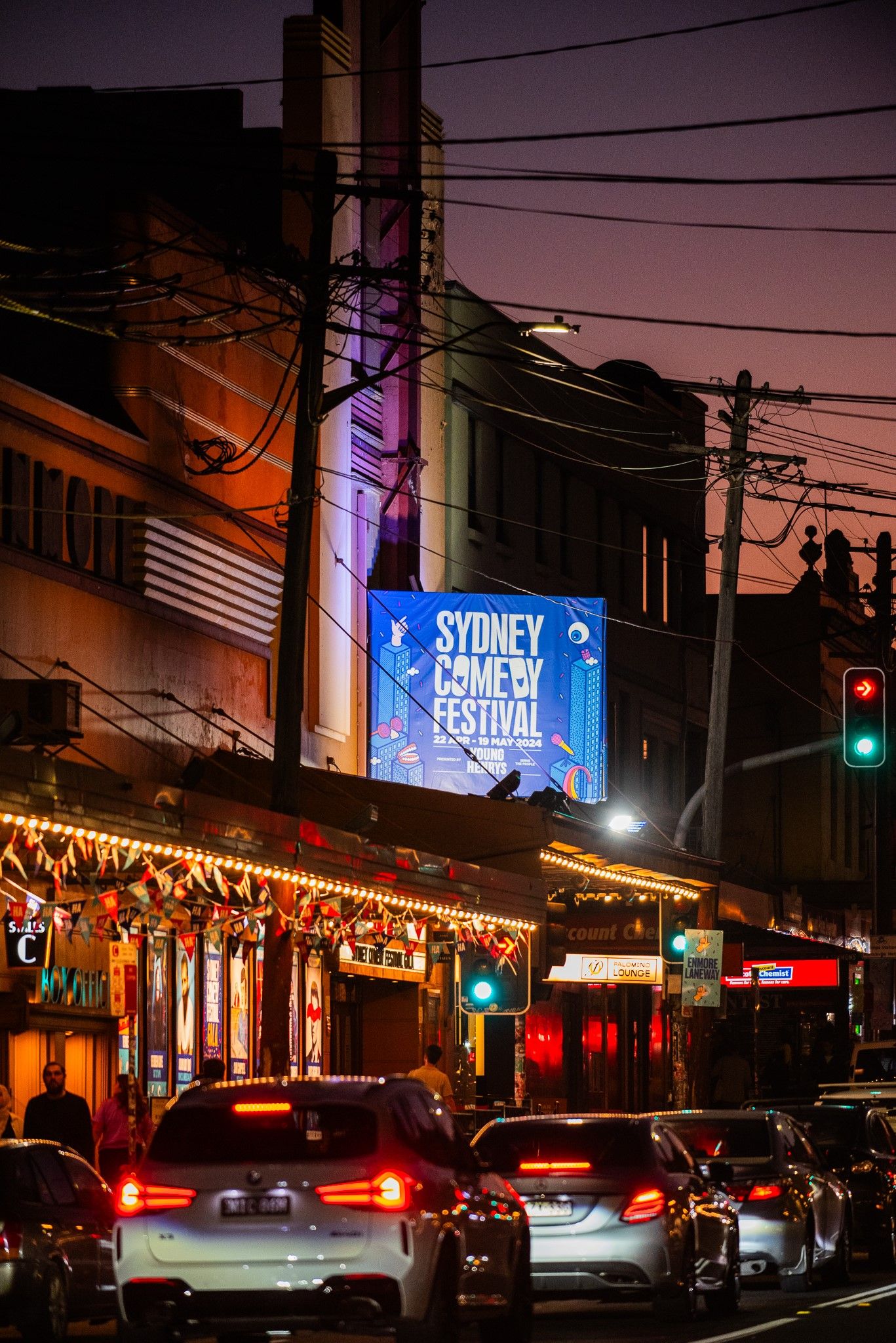 Street scene at dusk with traffic, shops, and a lit sign for the Sydney Comedy Festival above.