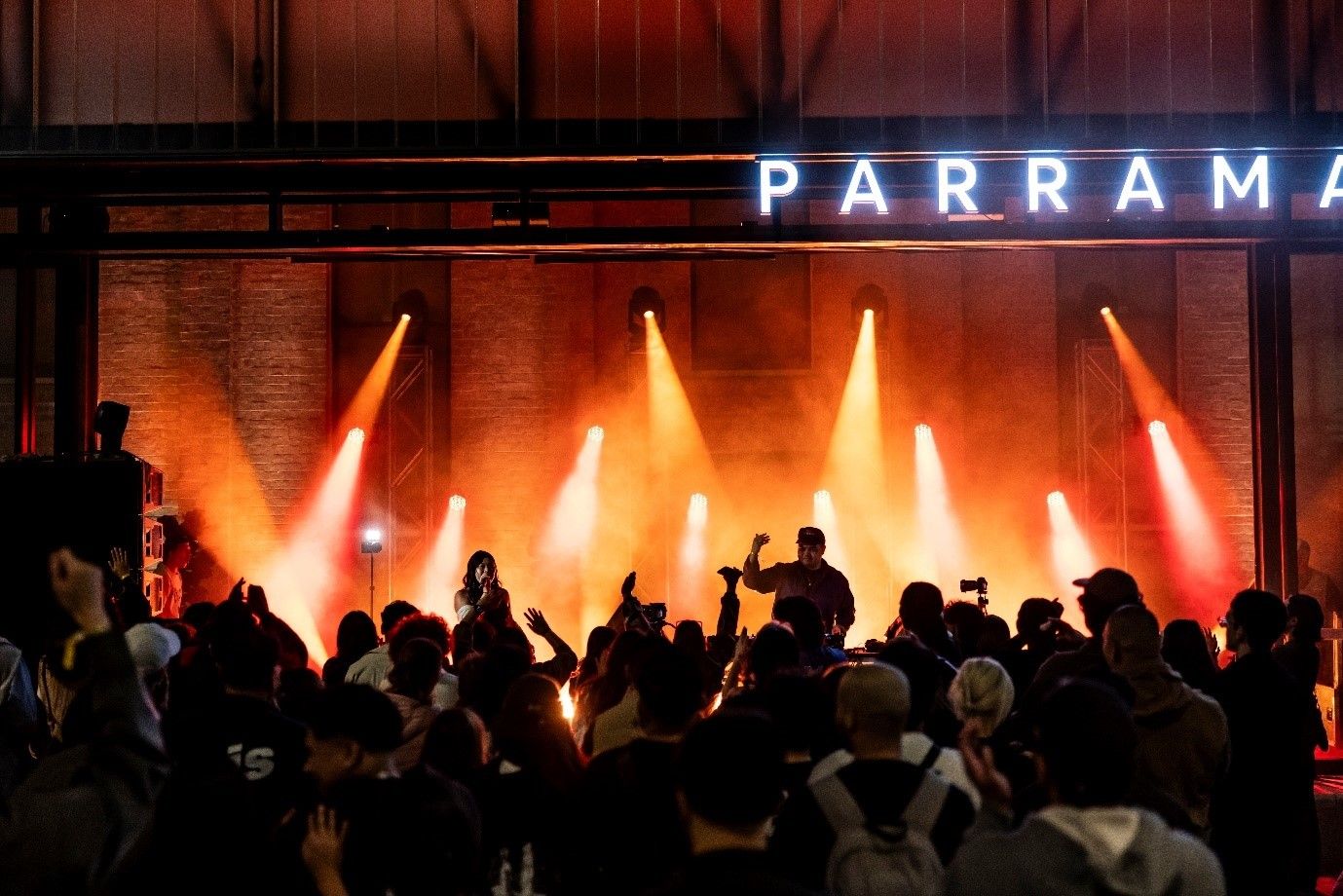 Performers on stage, with orange lighting at night, with a crowd of people in front of them