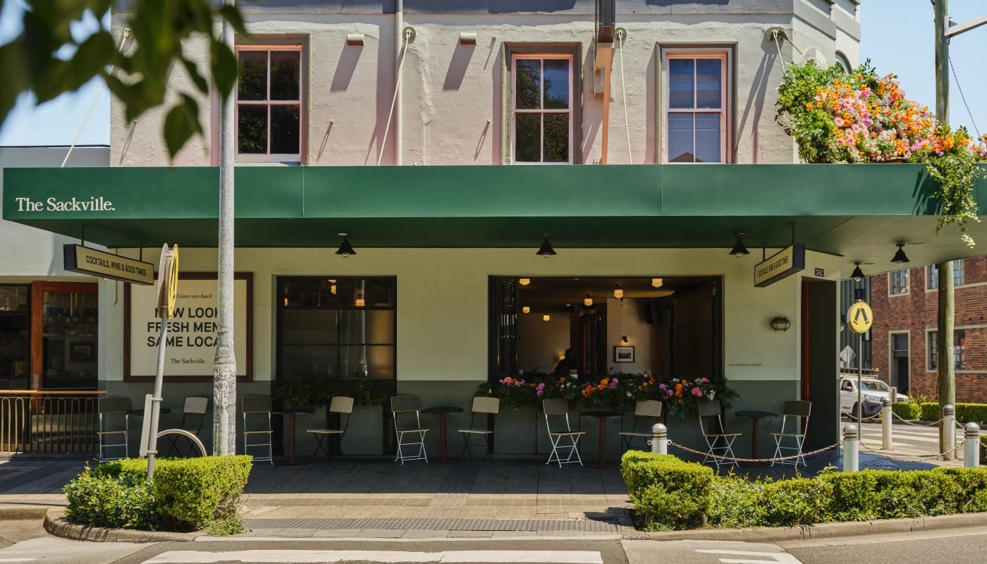 Exterior of a café with a green awning and potted flowers, featuring outdoor seating on a sunny day.
