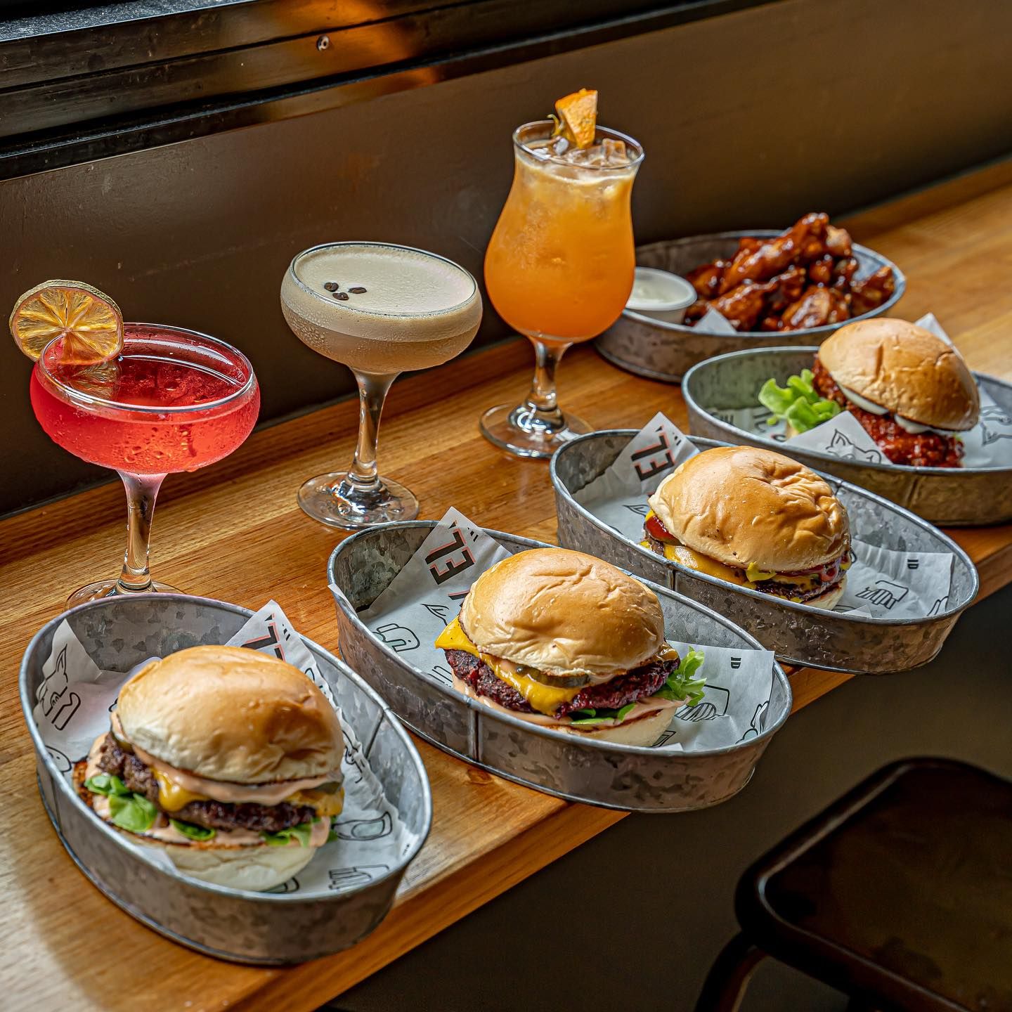 Burgers in metal trays with cocktails and chicken wings on a wooden table in a restaurant setting.