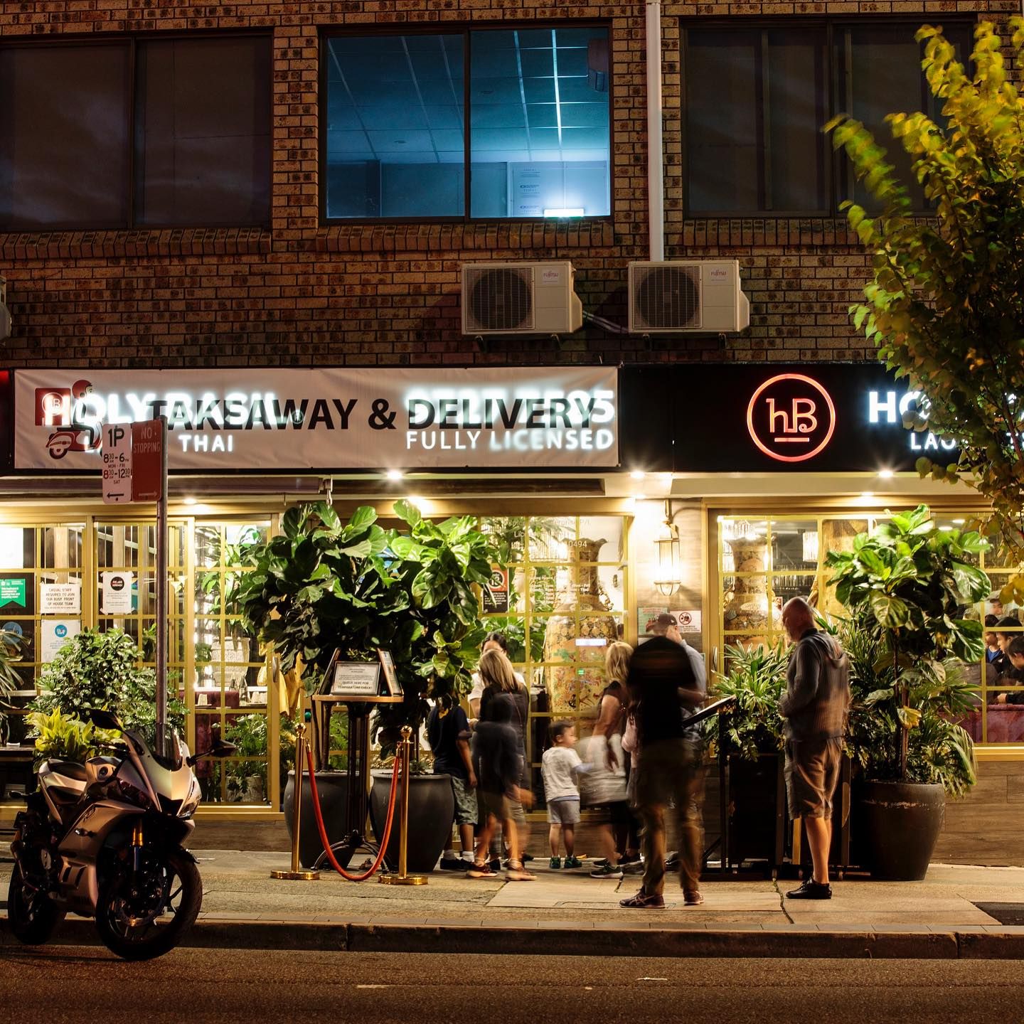 Night view of a lit restaurant entrance with people entering, a motorcycle parked outside, and large potted plants flanking the doorway.