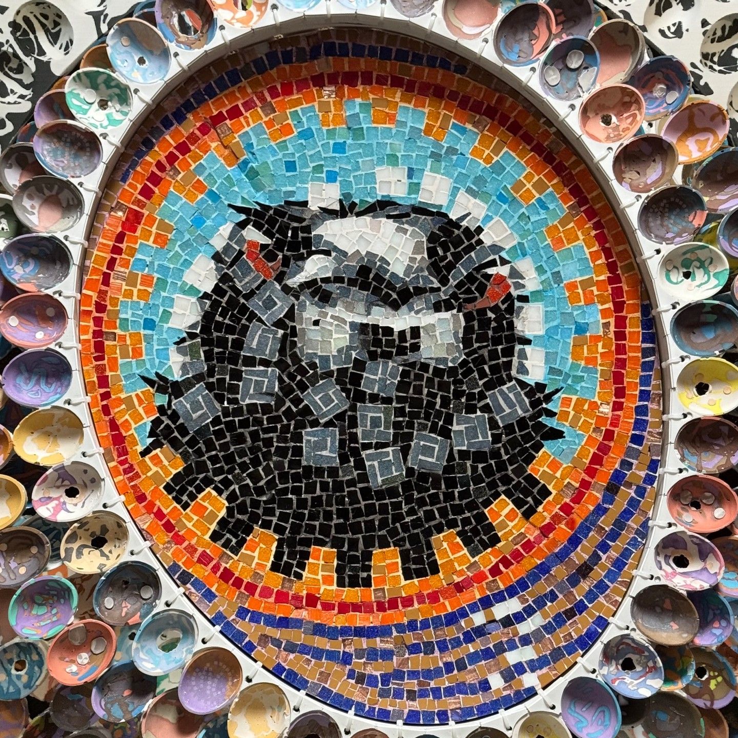 Colourful mosaic art with a fox motif in the center, surrounded by a circular frame of small, decorated cups.