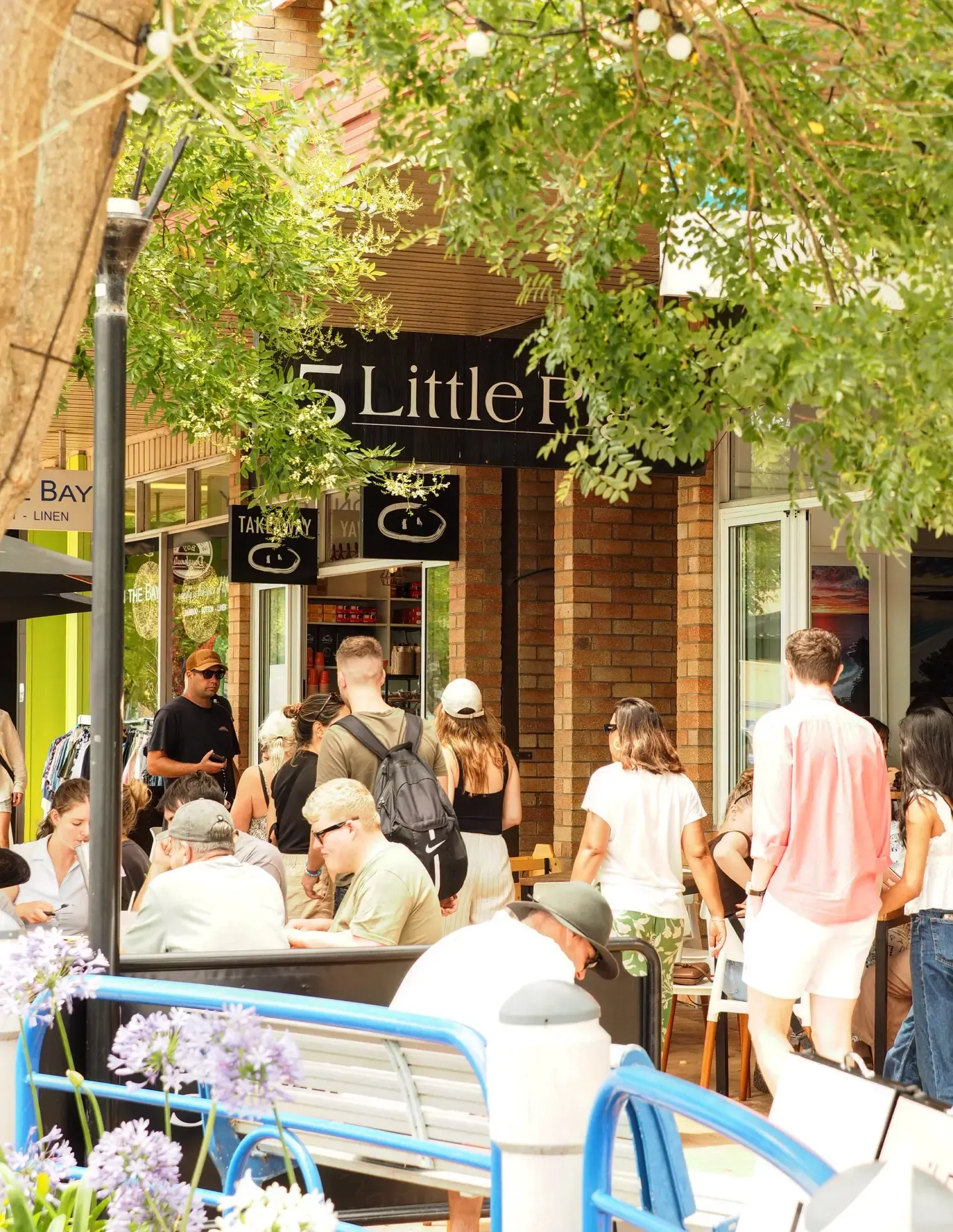 A bustling outdoor café scene with people dining and socializing beneath leafy trees next to a shop with a "5 Little" sign.