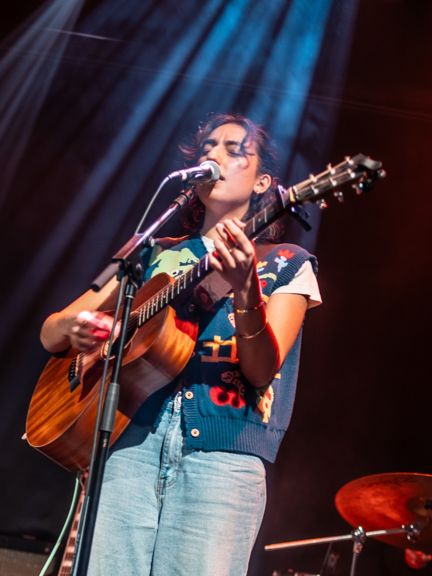 A person passionately sings and plays an acoustic guitar on stage, illuminated by colorful stage lights.