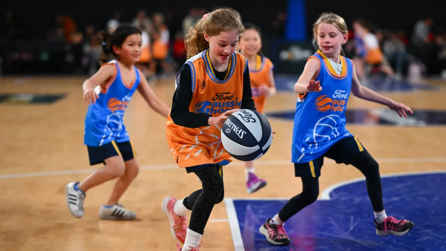 Young girls in blue and orange jerseys play basketball on a court, focusing on the ball.