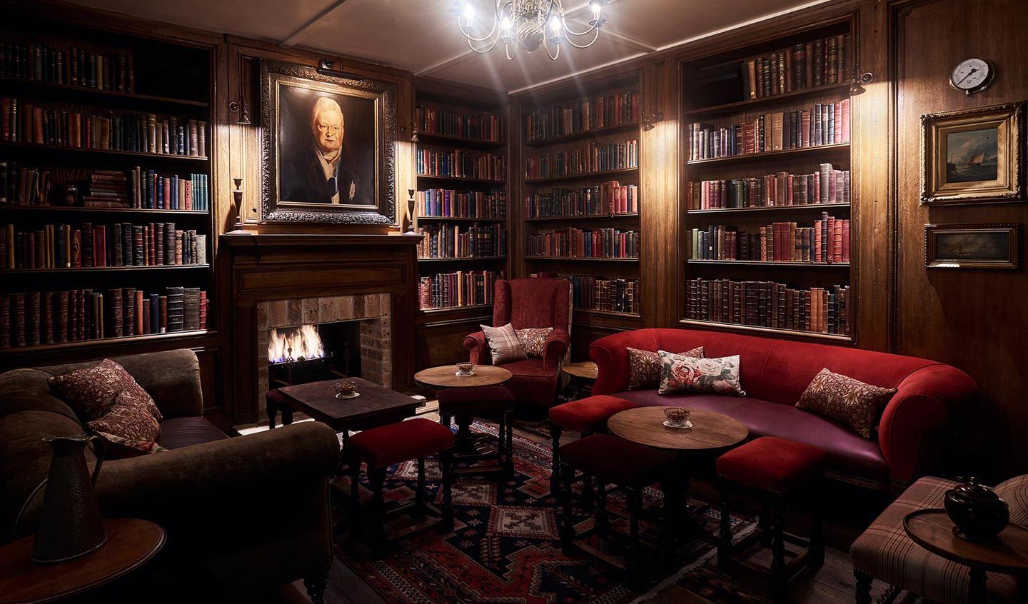 Cozy library with dim lighting, bookshelves lining the walls, red velvet seating, a warm fireplace, and a framed portrait above the mantel.