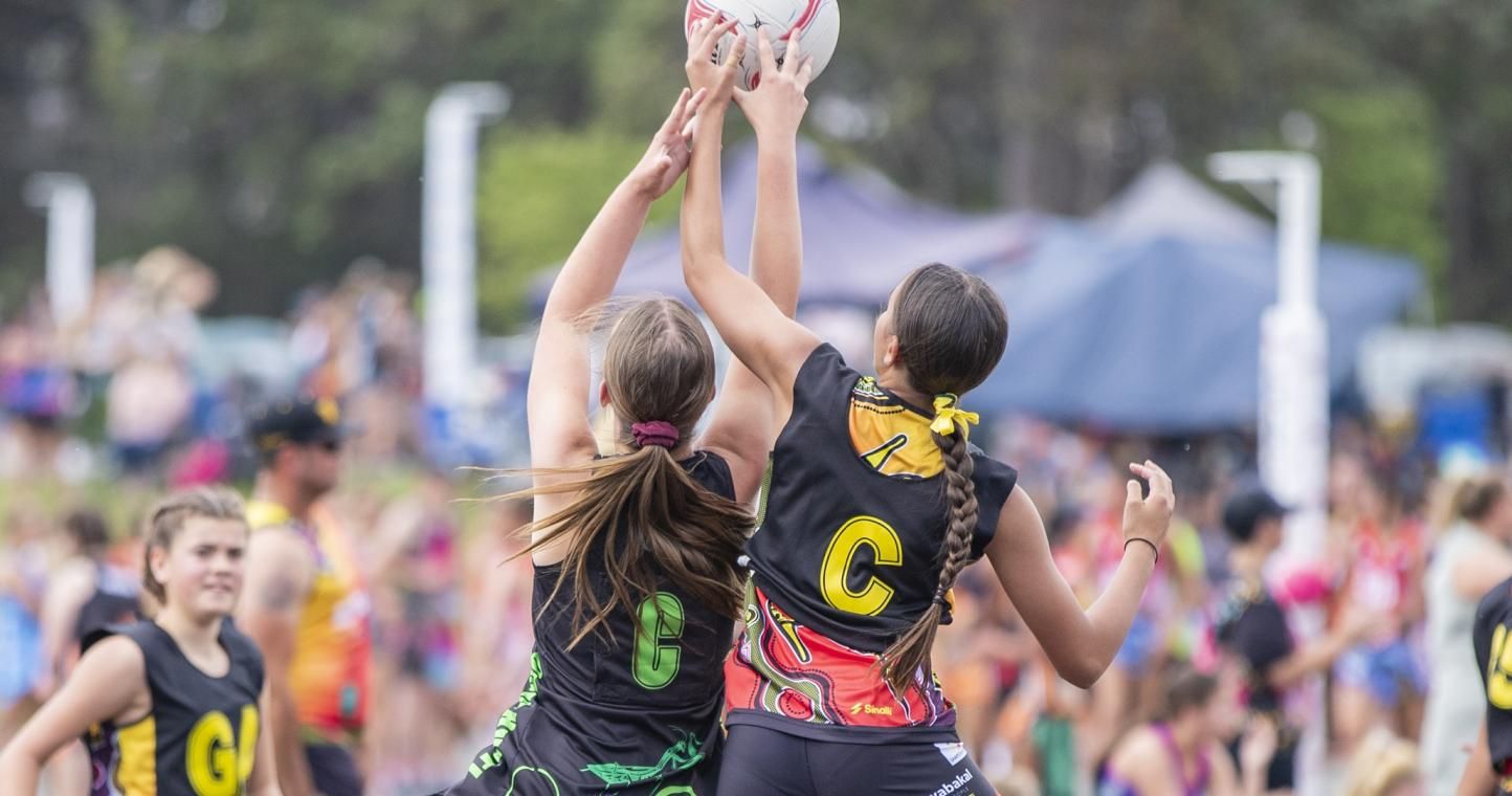 Two netball players jump for the ball during a match, with a blurred outdoor crowd and greenery in the background.