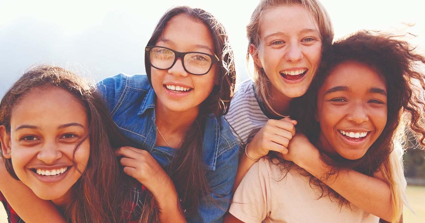 Four friends smiling and laughing outdoors, with one giving a piggyback ride. Sunlight creates a warm, cheerful atmosphere.