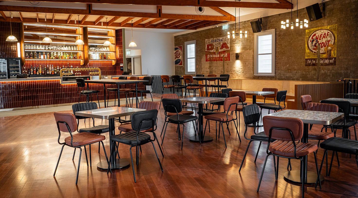 A cozy bar interior with empty tables and chairs on wooden flooring, a stocked bar, vintage decor, and warm lighting.