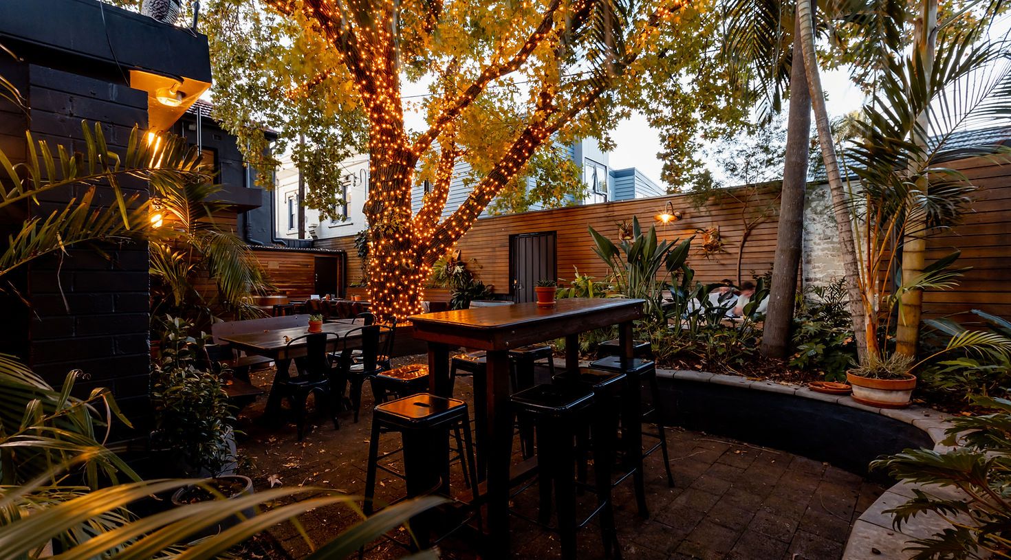 Outdoor patio with a tall table and chairs, surrounded by lush plants and a large tree wrapped in string lights, creating a cozy, warm atmosphere.