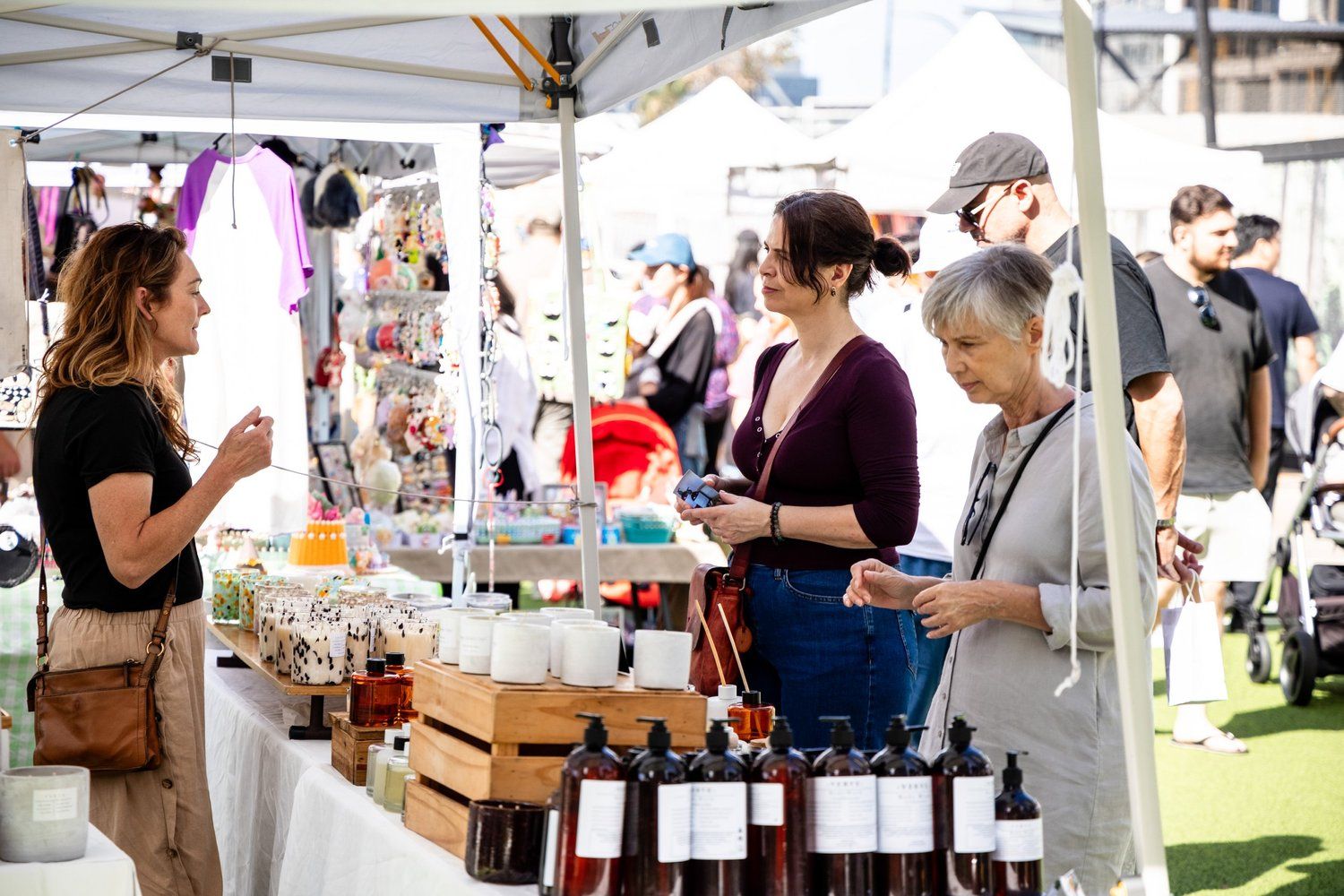 People browse skincare and home products at an outdoor market under a white tent. Various items are displayed on tables.