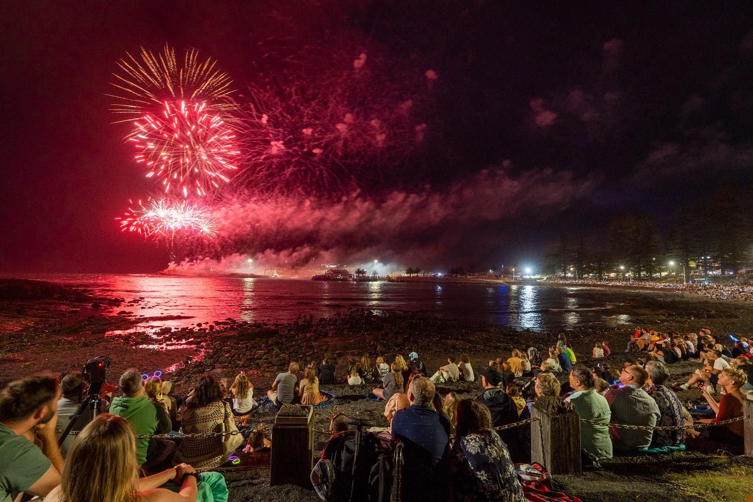 People sitting on a beach watching vibrant red fireworks illuminate the night sky over the water, with reflections and a distant lit-up shoreline.