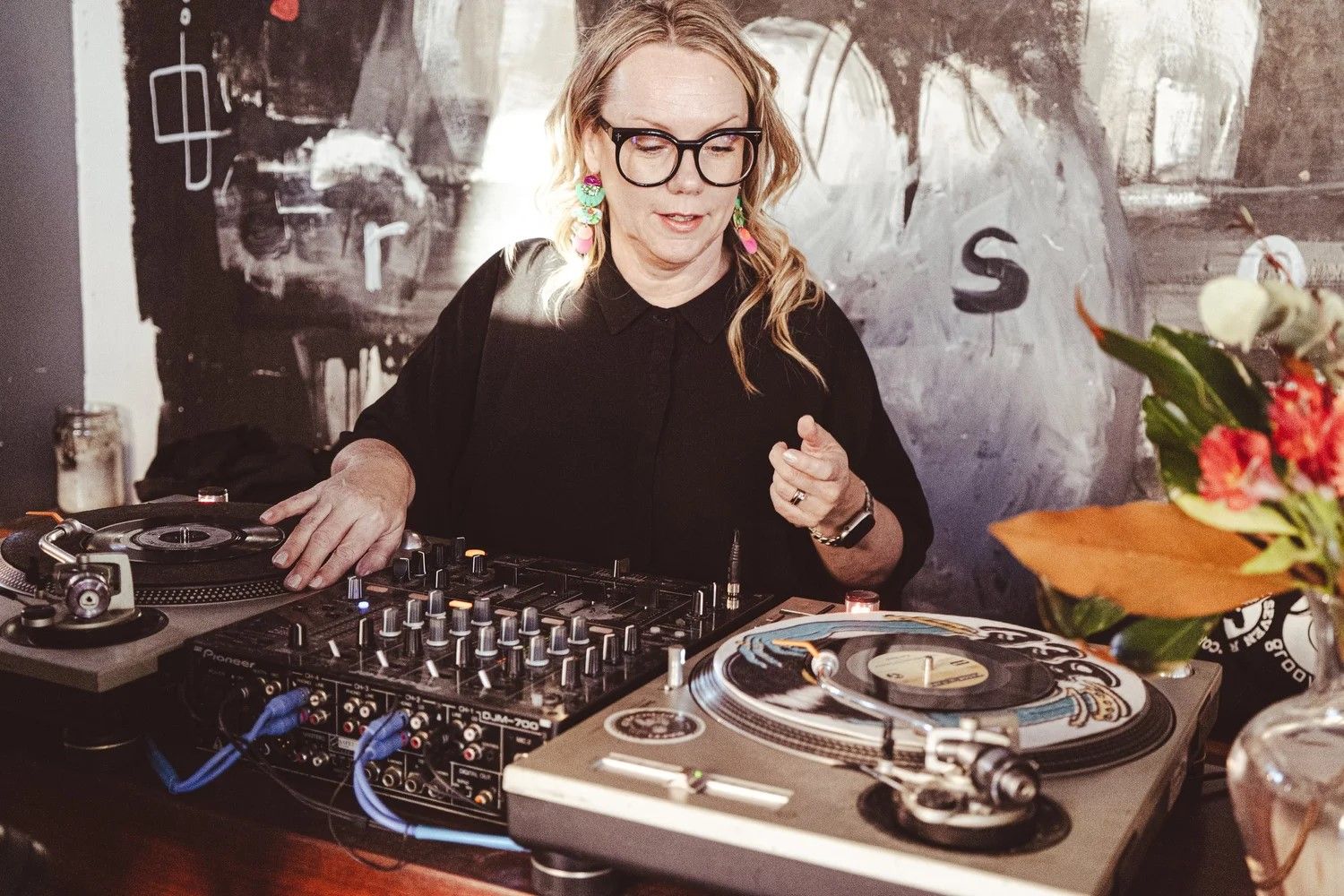 Woman DJ with glasses mixes vinyl records on turntables. Colorful earrings and a vibrant mural in the background add artistic flair.