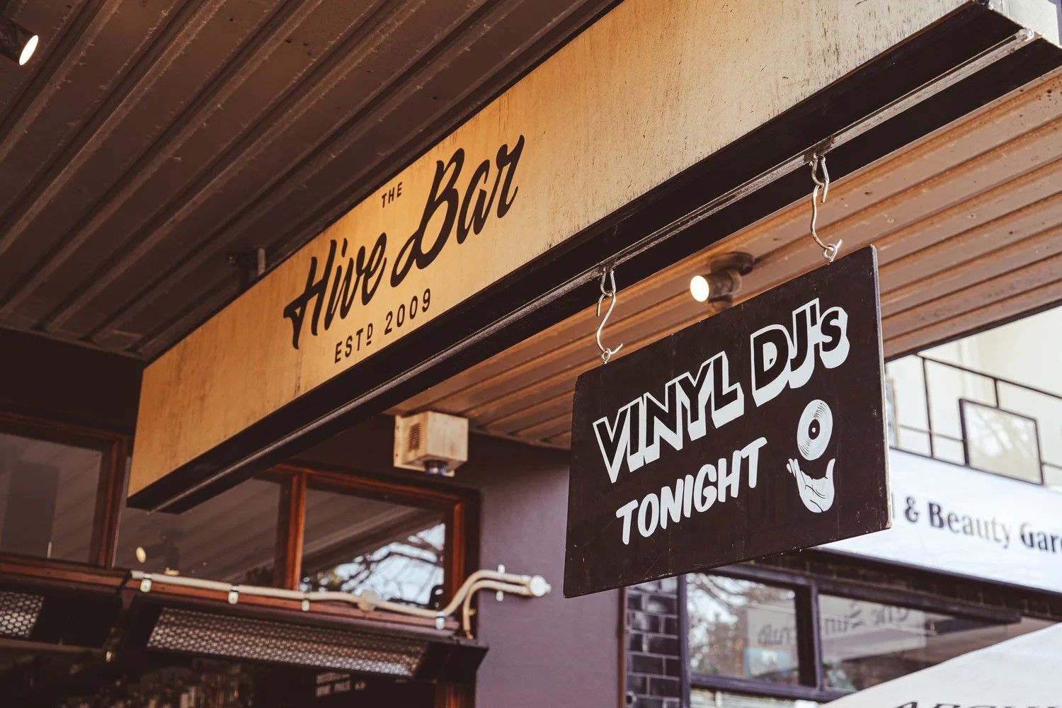Street view of "The Hive Bar" with a sign advertising "Vinyl DJ's Tonight." The bar has a wooden awning and glass windows.