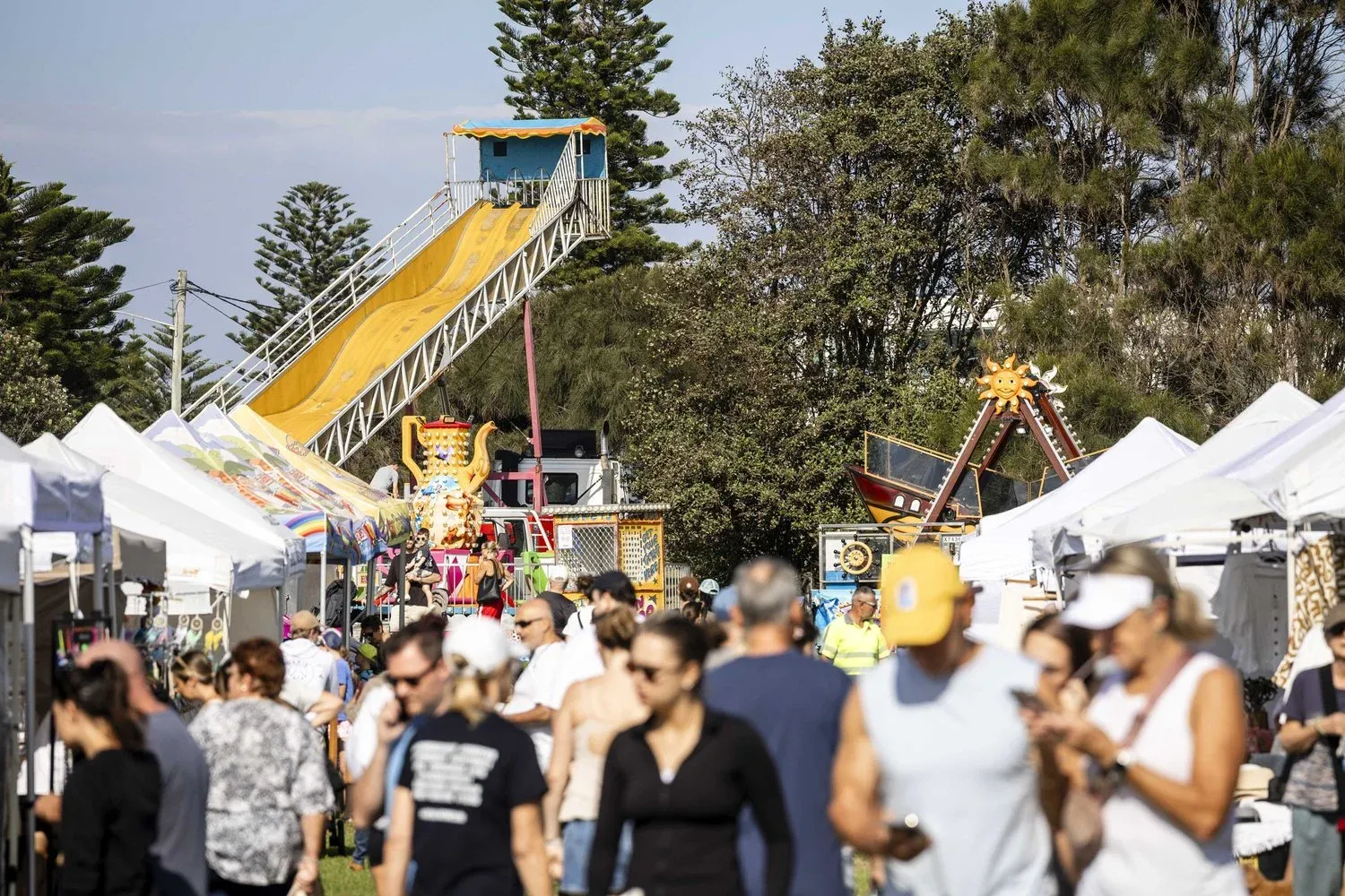 People stroll through a lively outdoor festival with white tents, a large slide, and trees in the background under a clear sky.