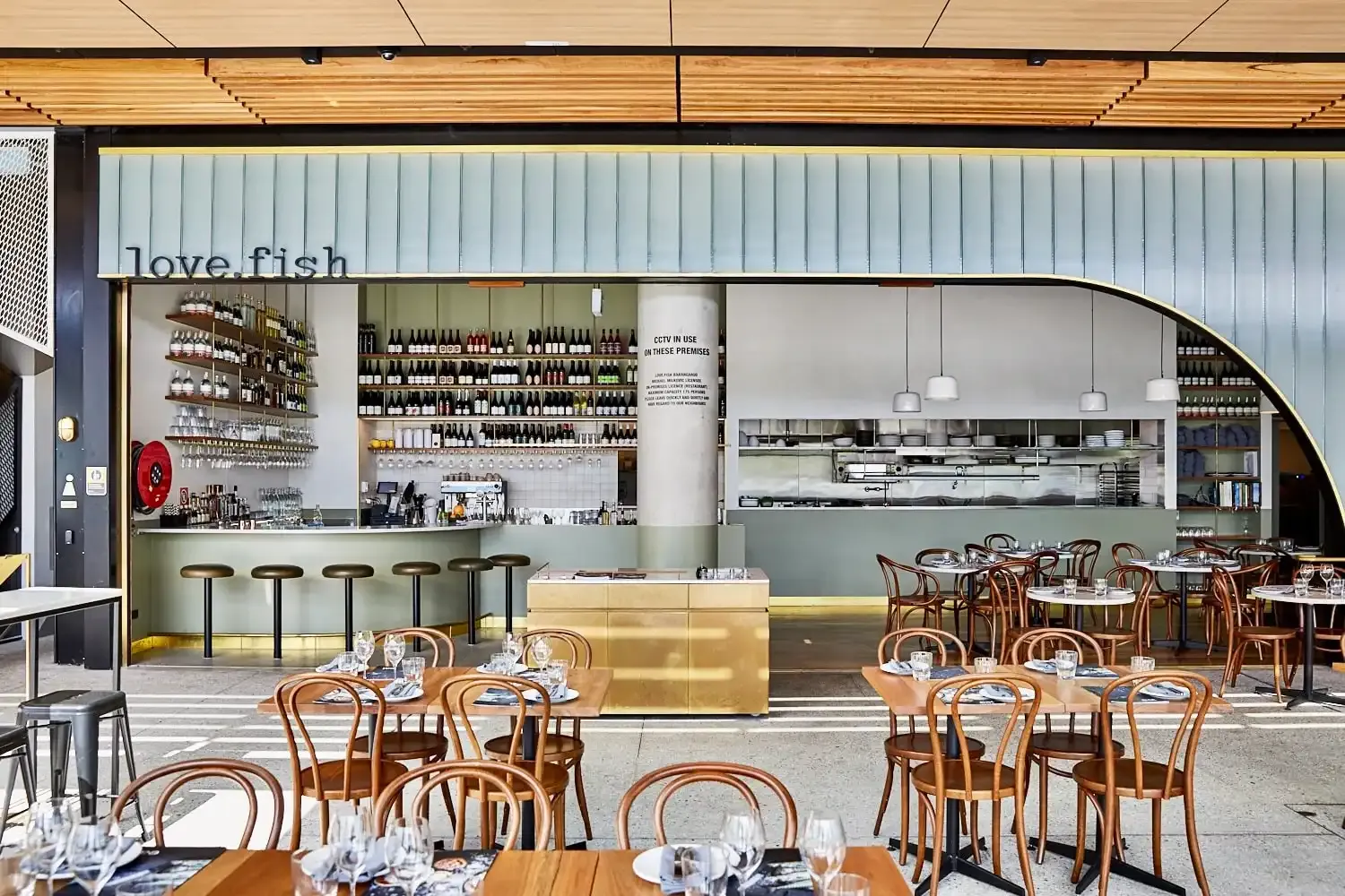 Modern restaurant interior with wooden chairs, white tables, and a bar with wine bottles displayed. The restaurant name, "love.fish," is visible.