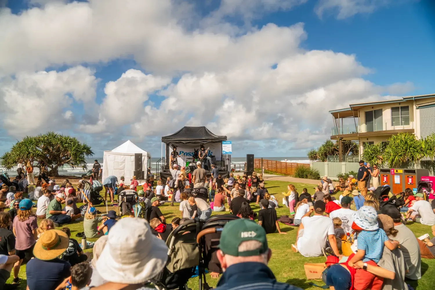 A lively outdoor music event with a diverse crowd seated on grass, enjoying a performance on a small stage under a bright, partly cloudy sky.