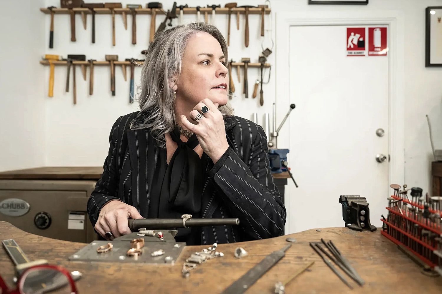 Person with gray hair in a striped suit sits at a jewelry workbench, surrounded by tools and rings, looking to the side.