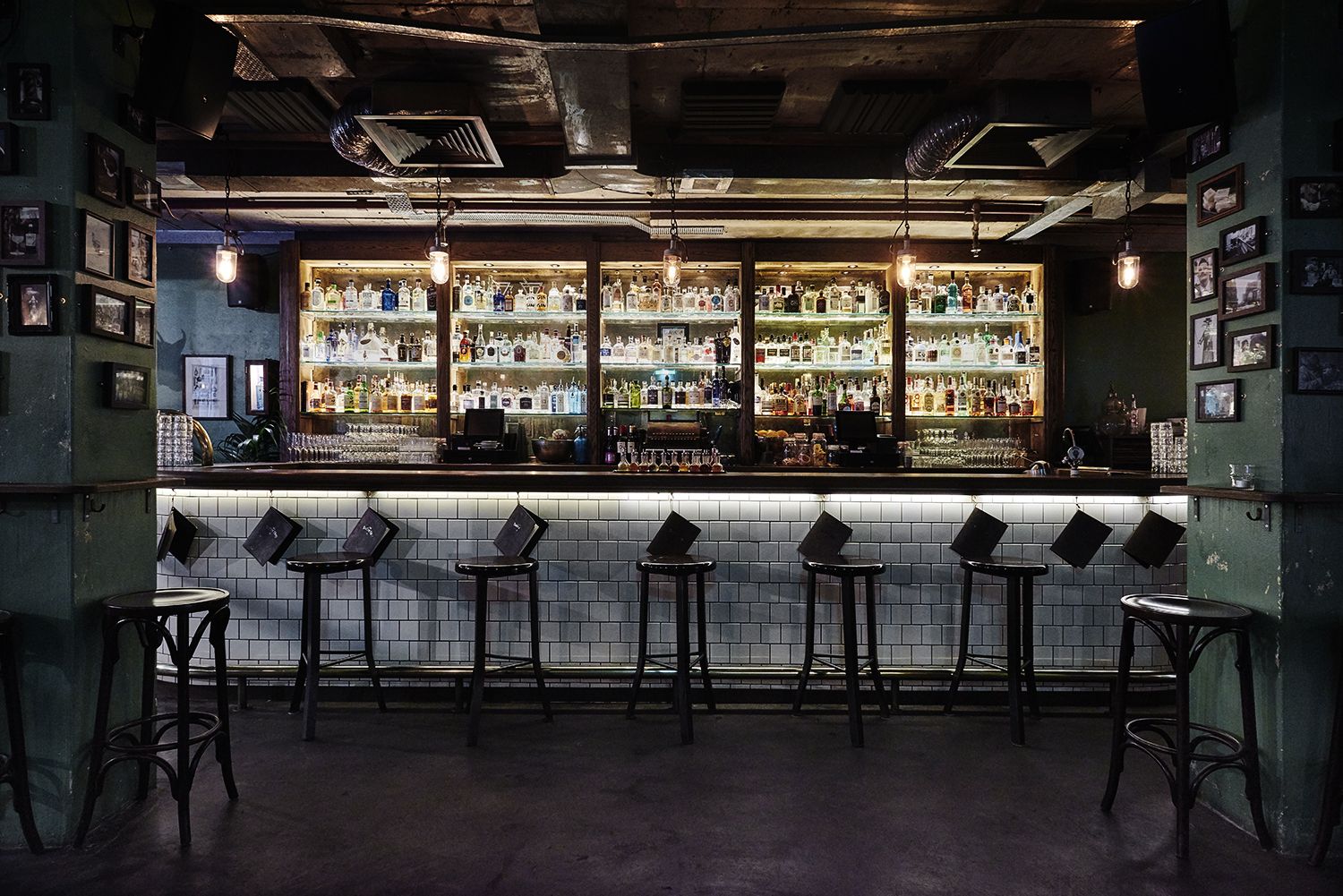 Dimly lit bar with a tiled counter, lined with high stools. Shelves behind display various bottles. Framed photos adorn the green walls.