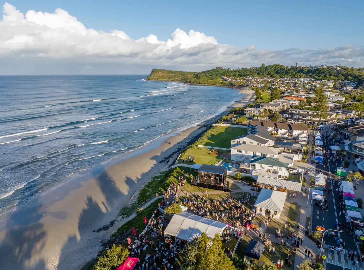 Coastal town with a festival along the beach, featuring tents and a stage. Waves roll onto the sandy shore with green hills in the background.