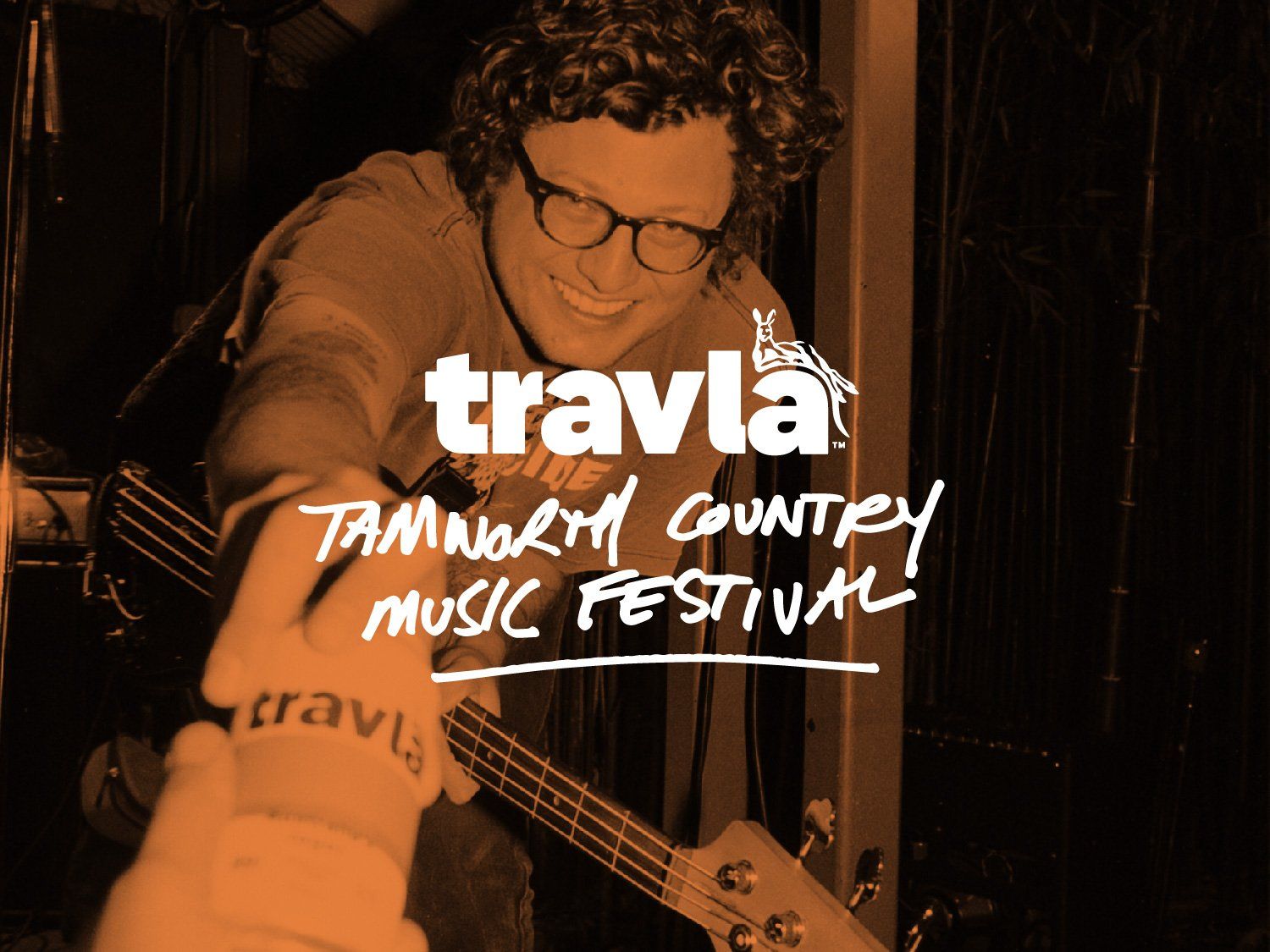 Musician with curly hair and glasses reaches to shake hands at the Tamworth Country Music Festival, with a guitar in the background.