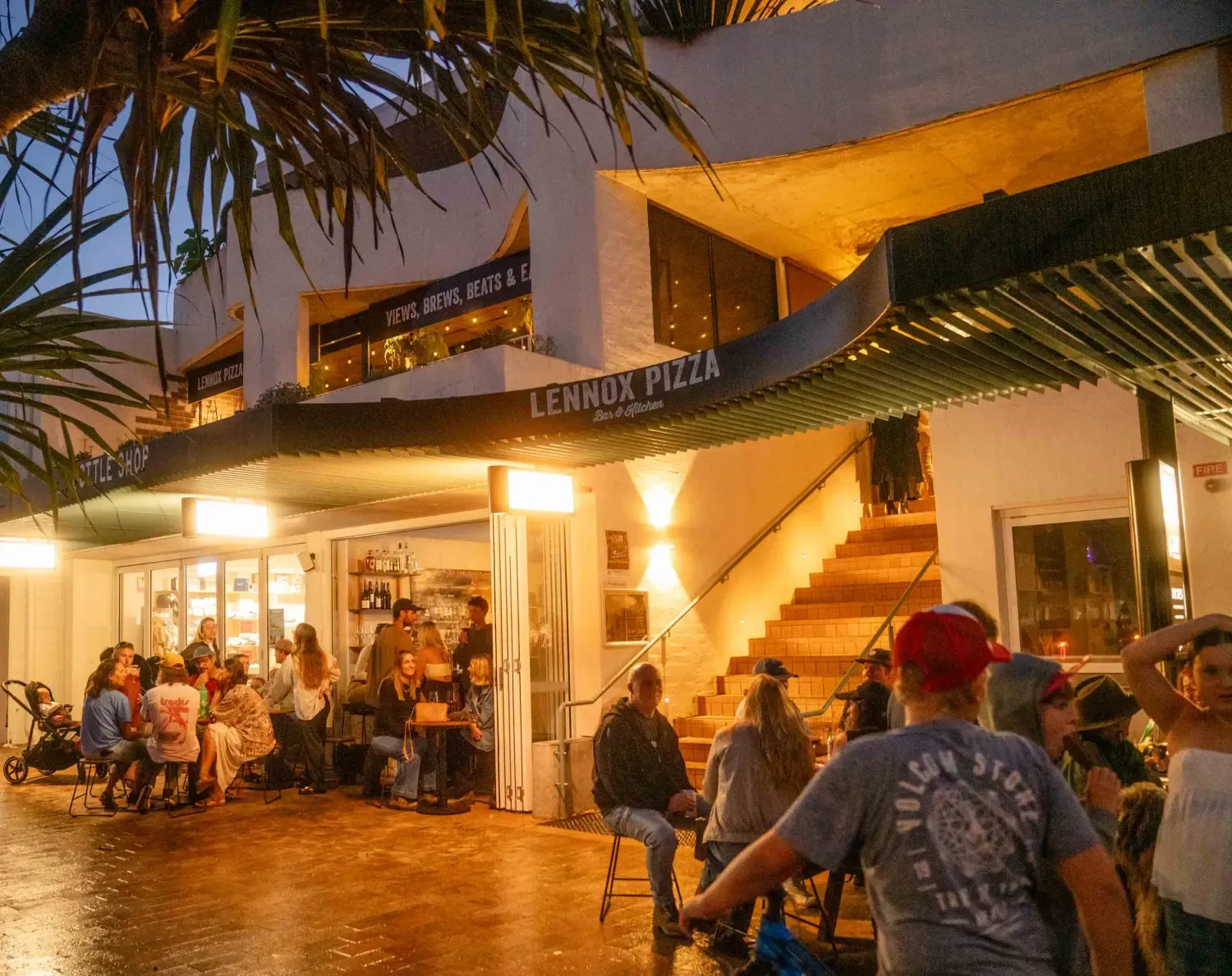 People gather outside Lennox Pizza in the evening, sitting and chatting under warm lights, with vibrant activity and a cozy atmosphere.