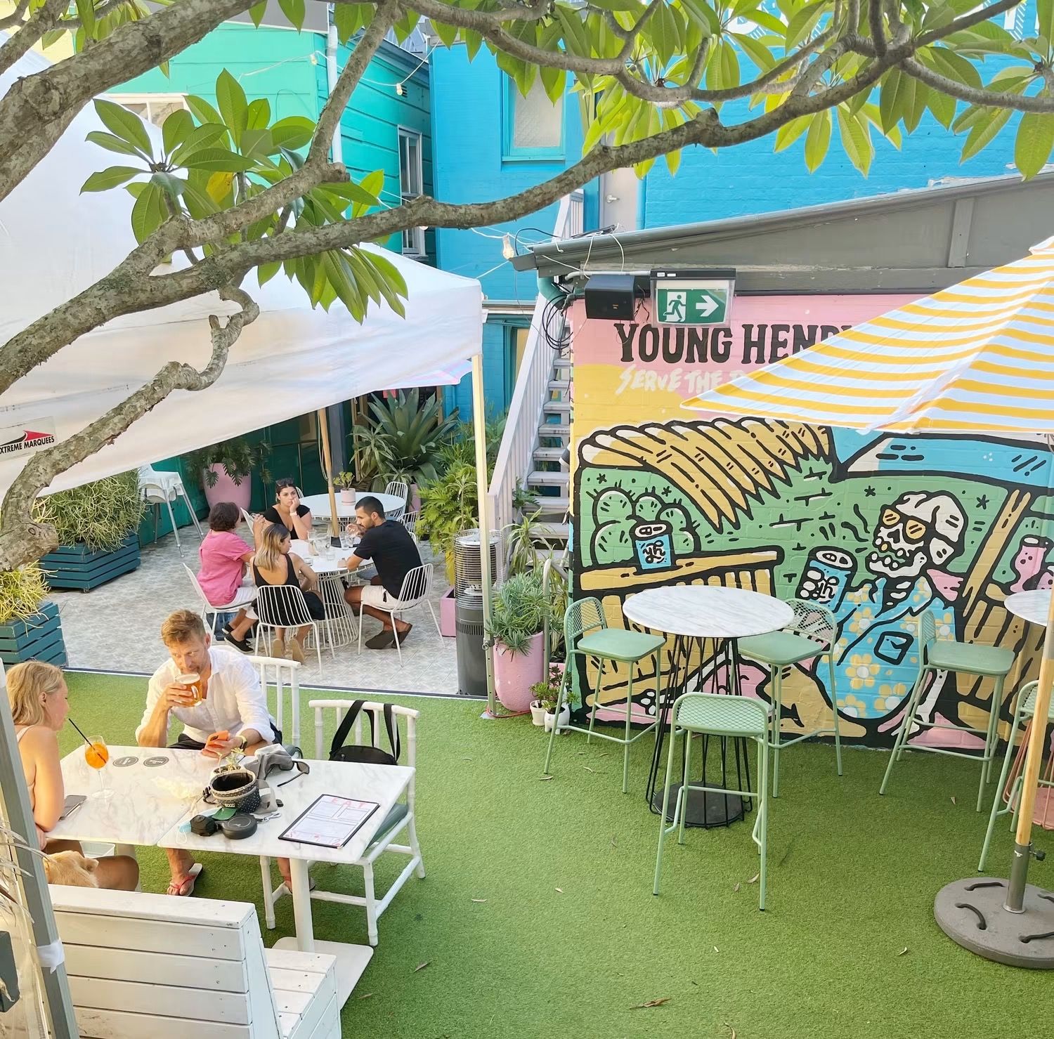 Outdoor cafe with people seated, colorful mural, green artificial grass, white tables, and striped umbrellas under a tree shade.