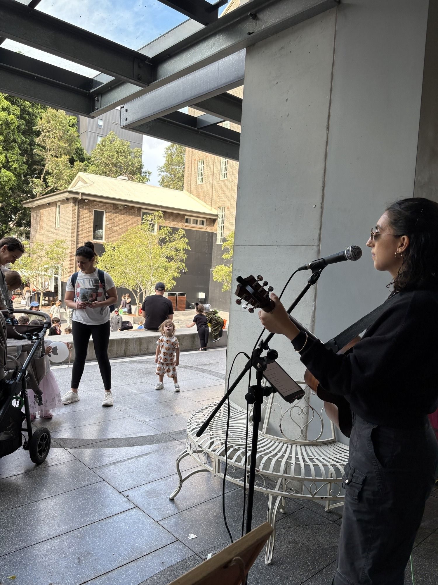 A musician performs with a guitar and microphone in an outdoor area, while people, including a woman with a stroller, pass by.