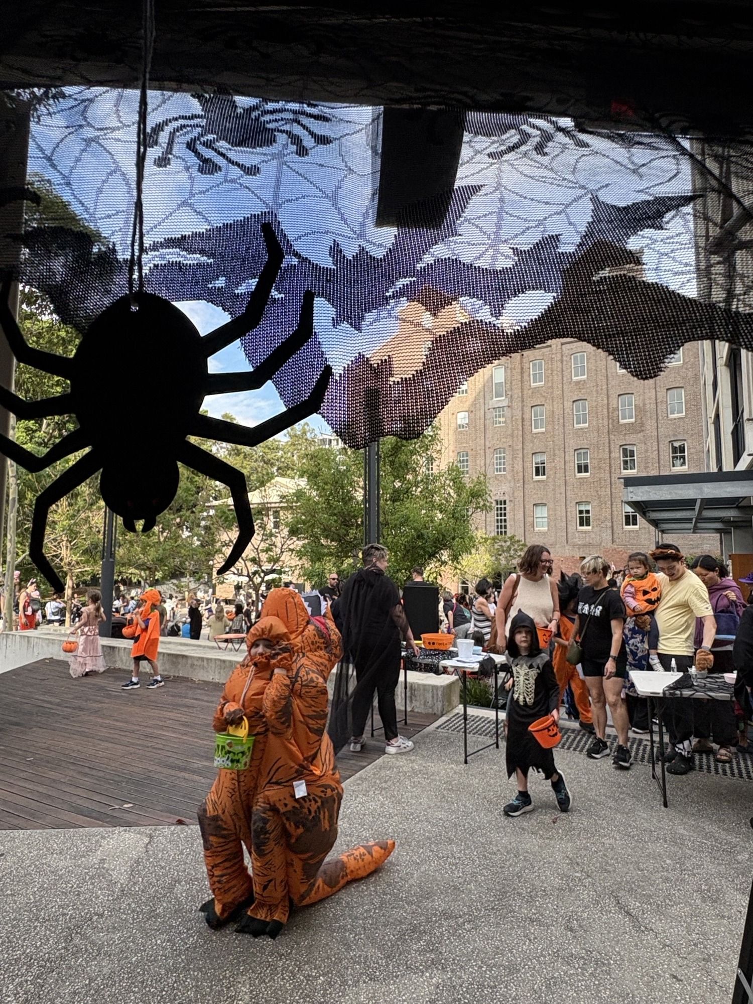 Halloween event with people in costumes, including tigers and pumpkins, under a large spider decoration in an outdoor urban setting.