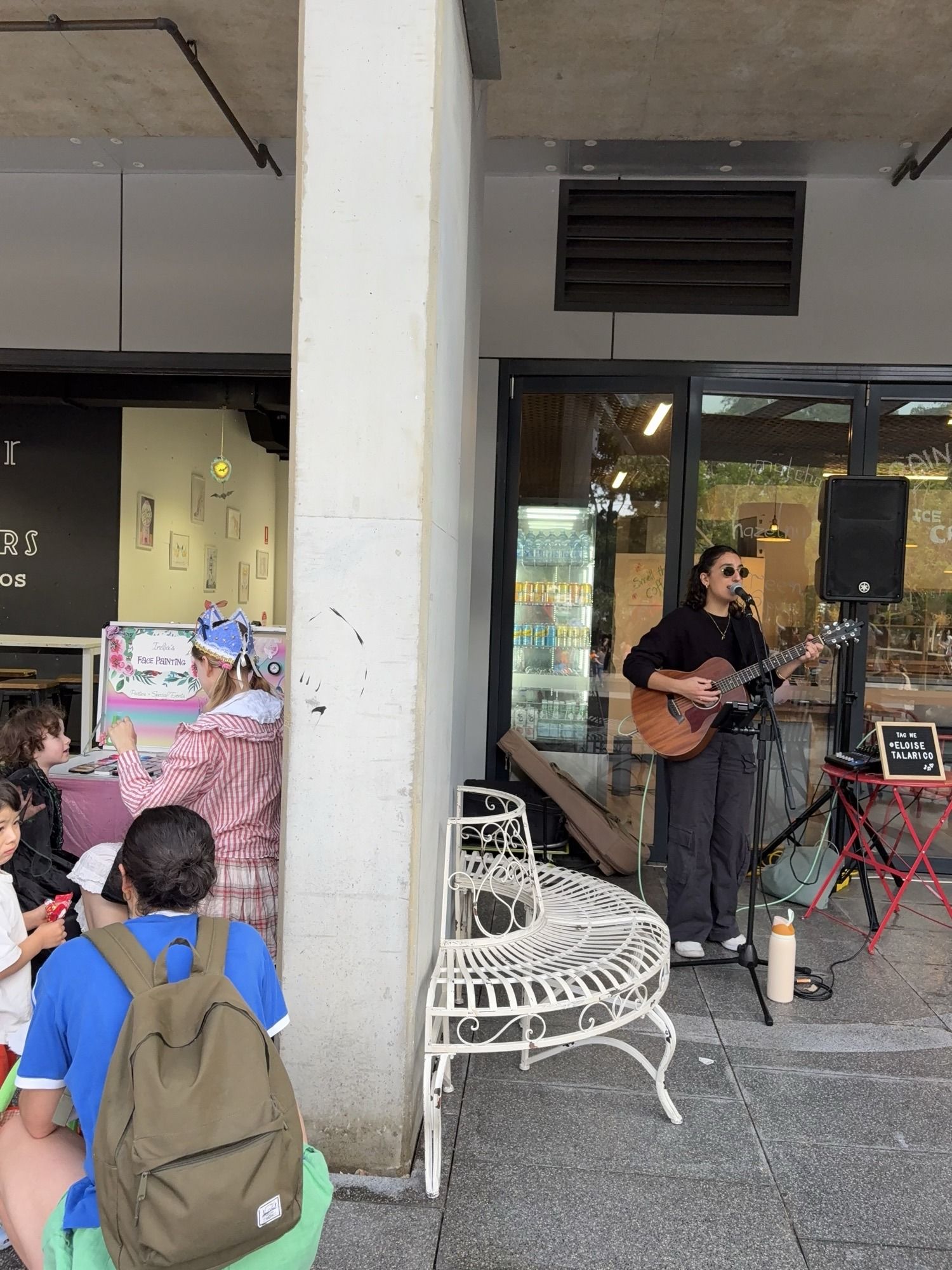 Musician playing guitar and singing outside a café, with people sitting nearby on benches and chairs.