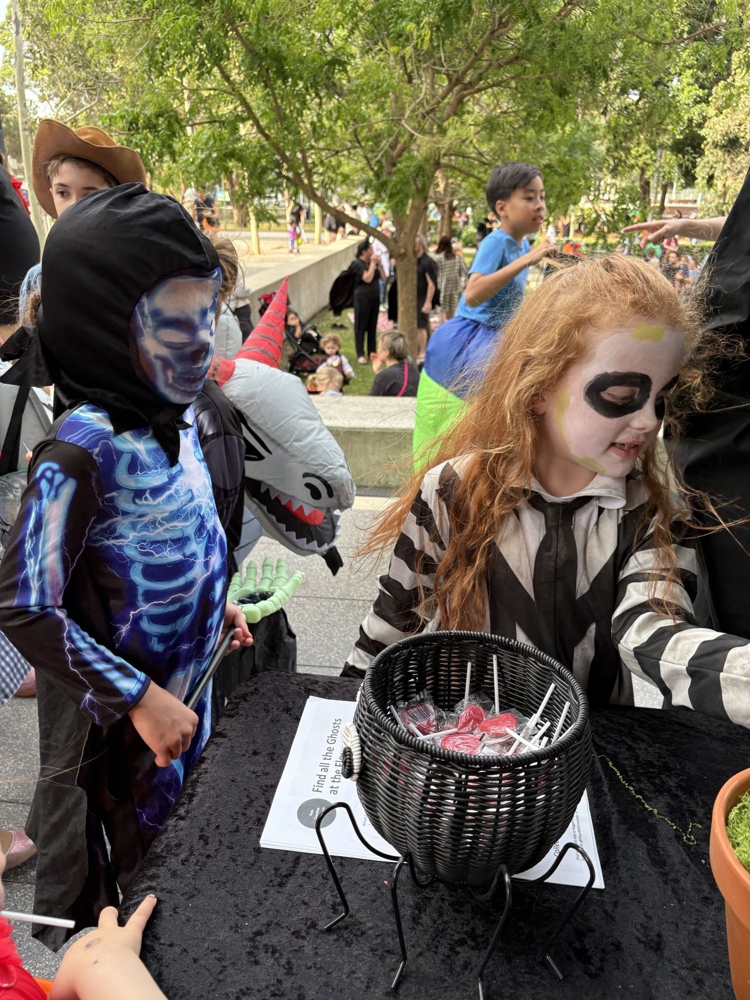 Children in Halloween costumes gather around a table with a black basket of lollipops. Trees and more people are visible in the background.