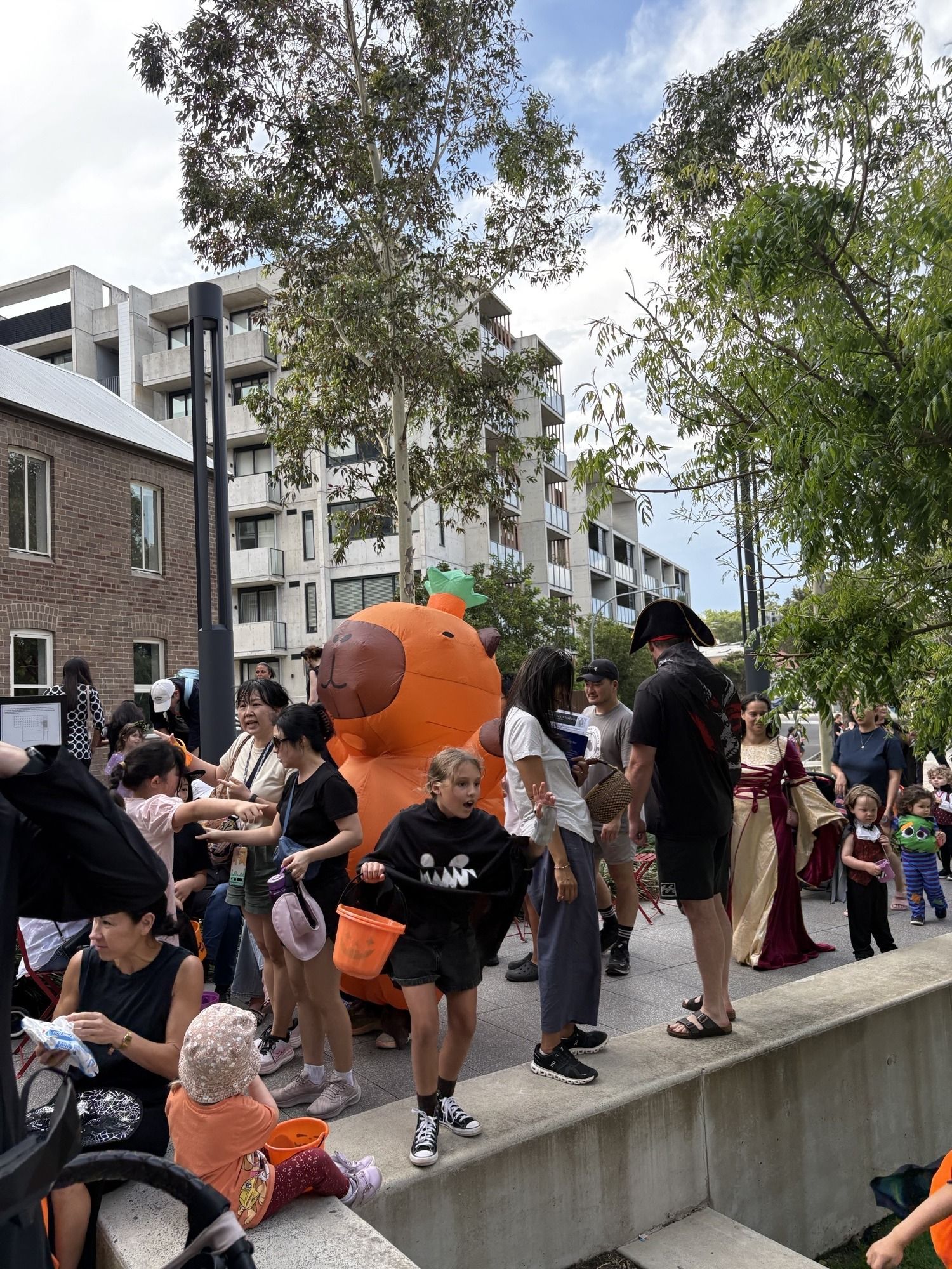 People in costumes at an outdoor event, including a large, inflatable pumpkin costume. Trees and buildings are in the background.