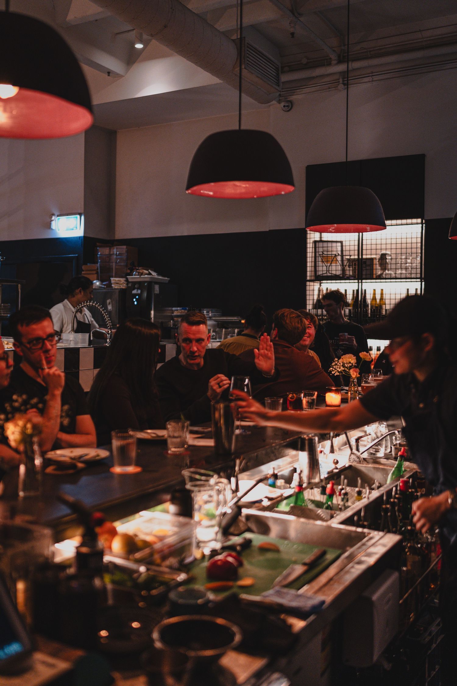 Dimly lit bar with people socializing and a bartender serving drinks. Hanging lamps cast a warm glow over the counter with drinks and garnishes.