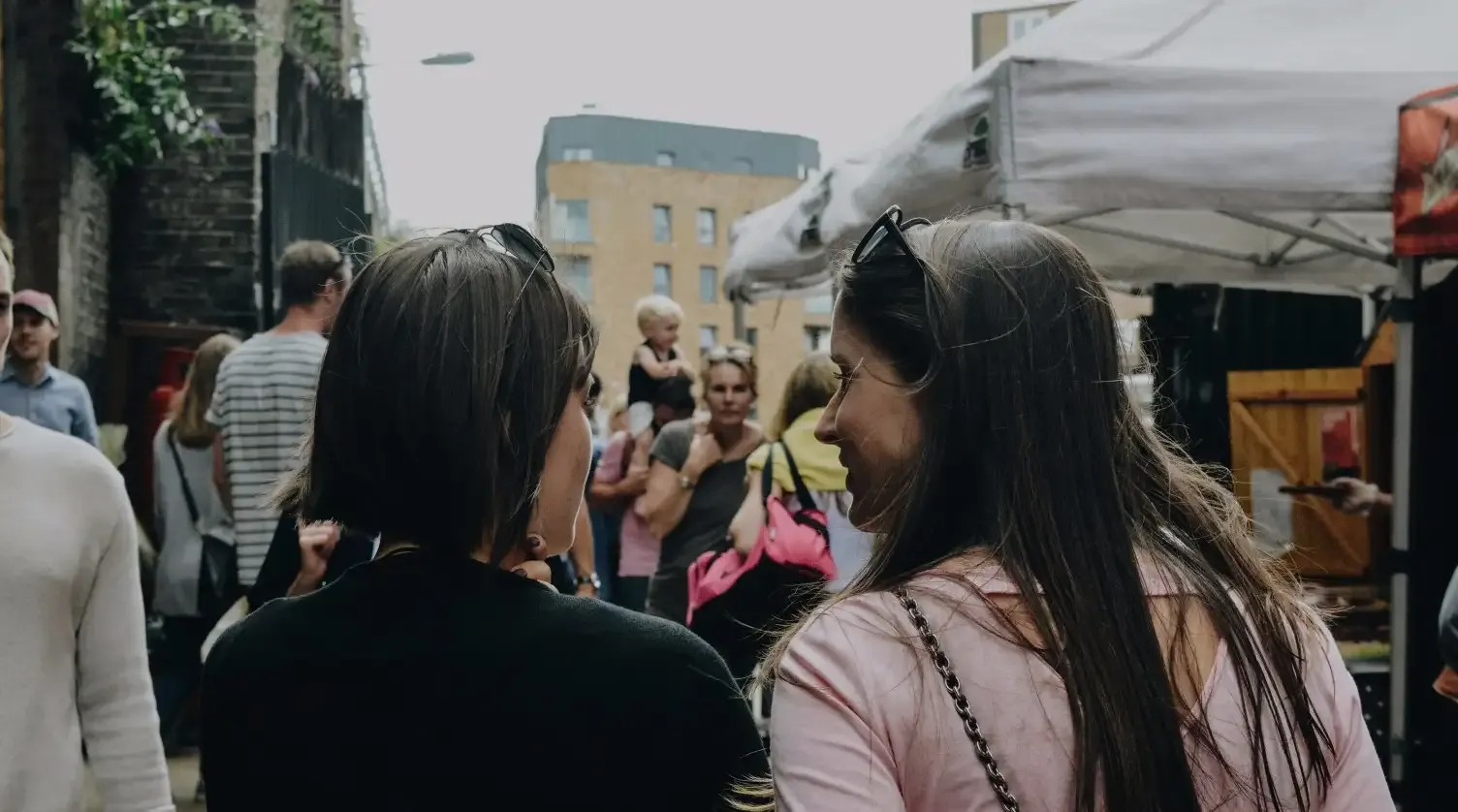 Two women walking and chatting in a bustling outdoor market, surrounded by people, stalls, and a building in the background.