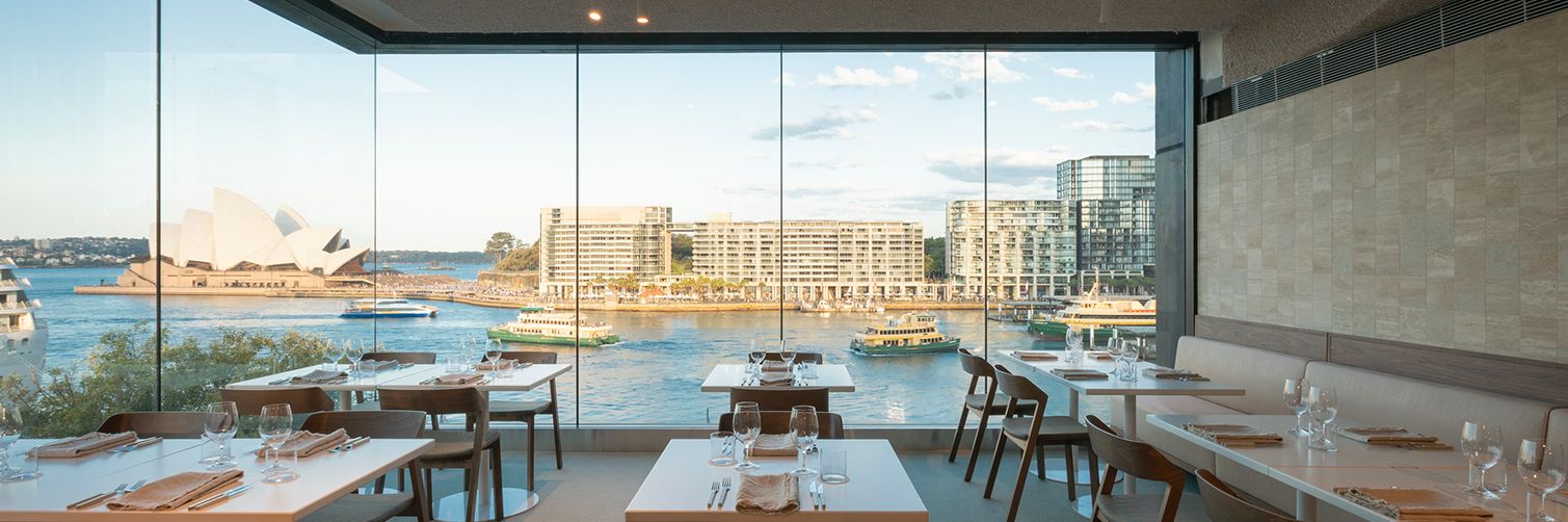 Elegant restaurant interior with tables set for dining; large window reveals a waterfront cityscape, including iconic architecture and boats.