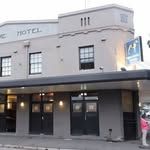 Two-story art deco style building with "Hotel" written on the front, featuring large windows and a blue neon sign on the side.
