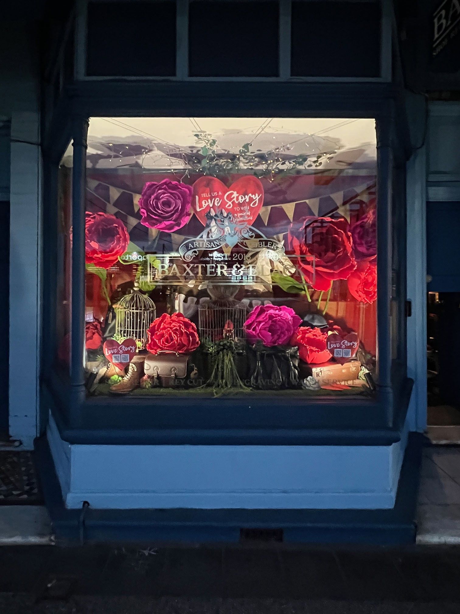 Storefront display with large red roses, birdcages, and a sign reading "The Greatest Love Story" and "Baxter & Ames."