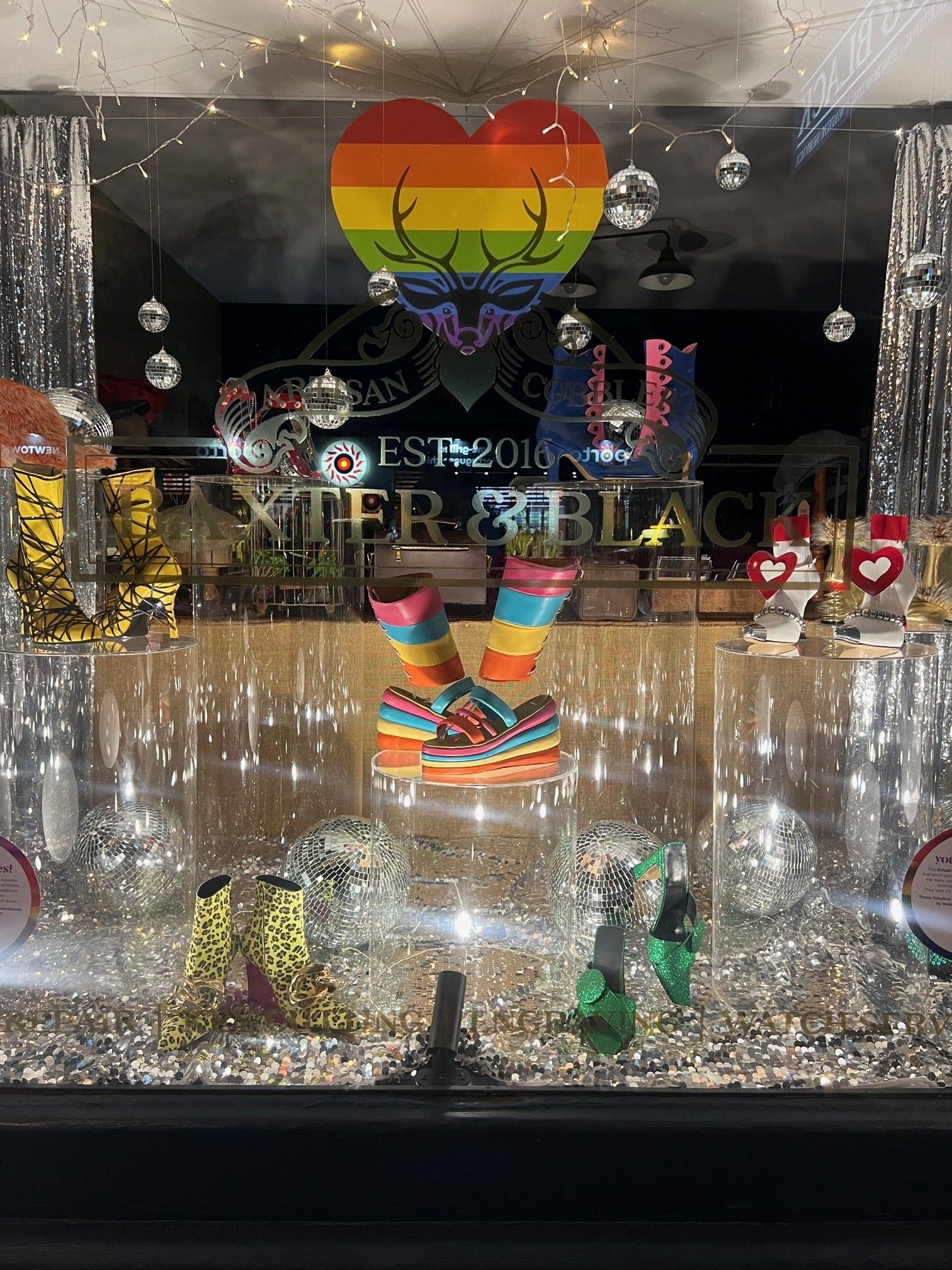 Storefront display with colourful platform shoes, disco balls, heart-shaped decorations, and a rainbow stag logo above.