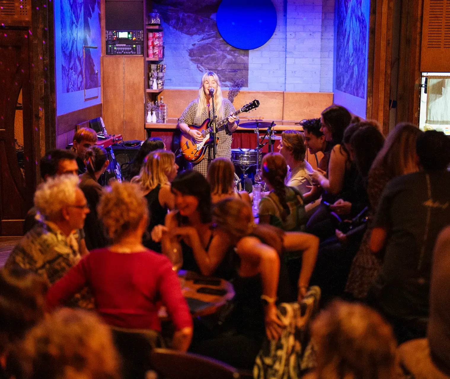 A musician with a guitar performs on stage in a cozy, dimly lit venue, surrounded by a crowd seated at tables.