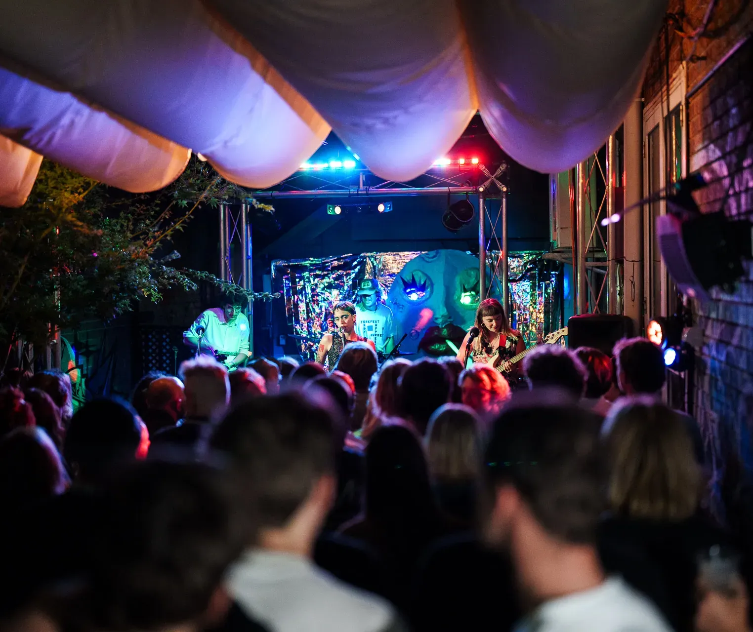 A band performing on an outdoor stage with colorful lights, surrounded by an attentive crowd under decorative fabric canopies.