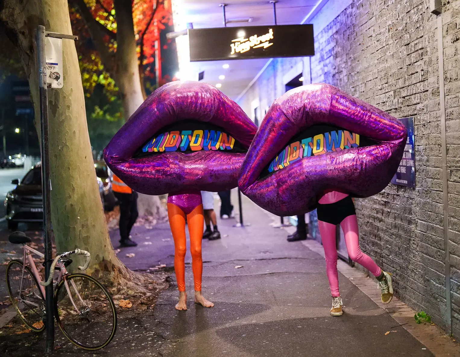 Two people in colorful leggings wear giant purple lip costumes reading "MIDTOWN" on a city street at night.
