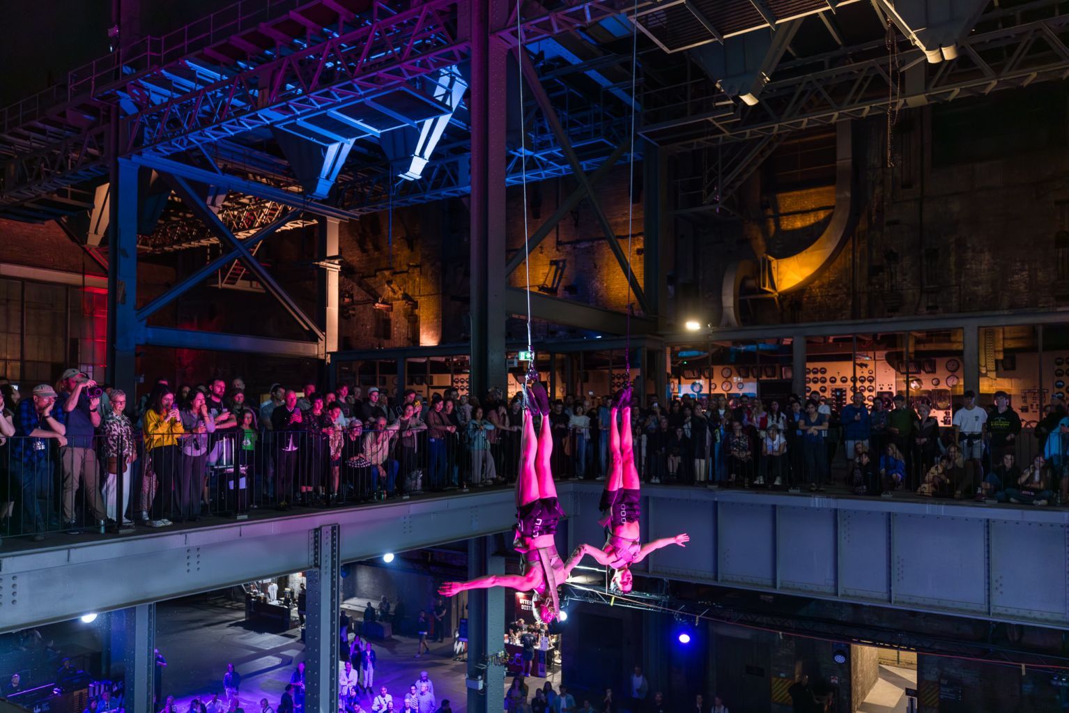 Two aerial performers suspended upside down with a crowd watching in an industrial-style venue with colorful lighting.