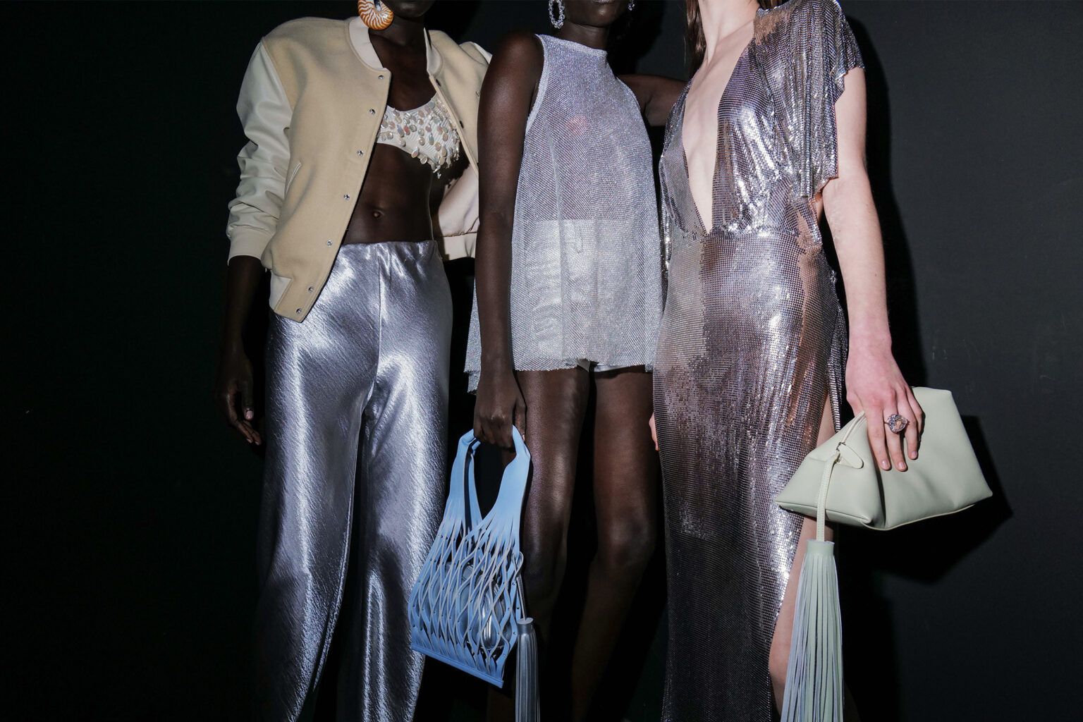 Three people in stylish metallic outfits, one holding a blue netted bag and another a cream clutch, pose against a dark backdrop.
