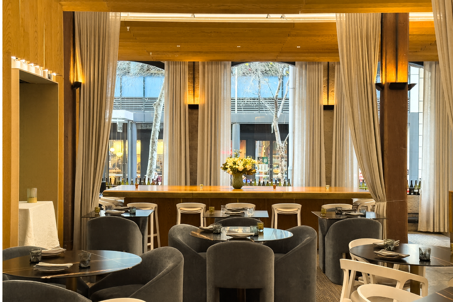 Elegant restaurant interior with round tables, cushioned chairs, and large windows with sheer curtains. A central bar features floral arrangements.