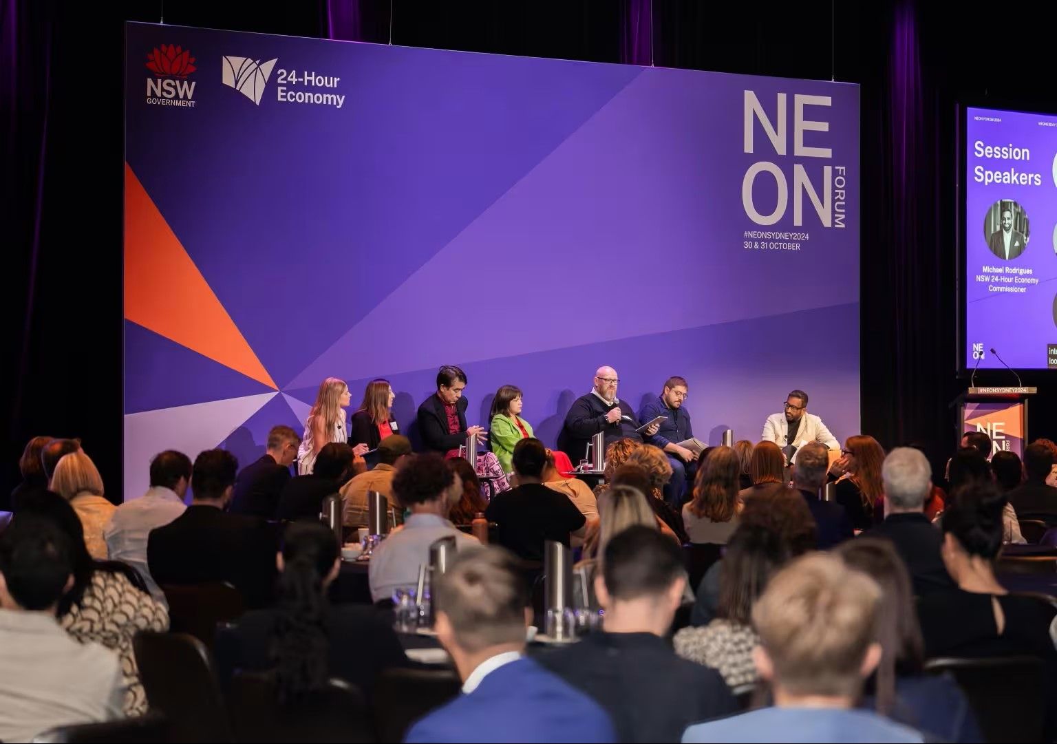 A panel of speakers sits on stage at a conference, with an audience watching. The backdrop displays "NEON Forum" and event details.