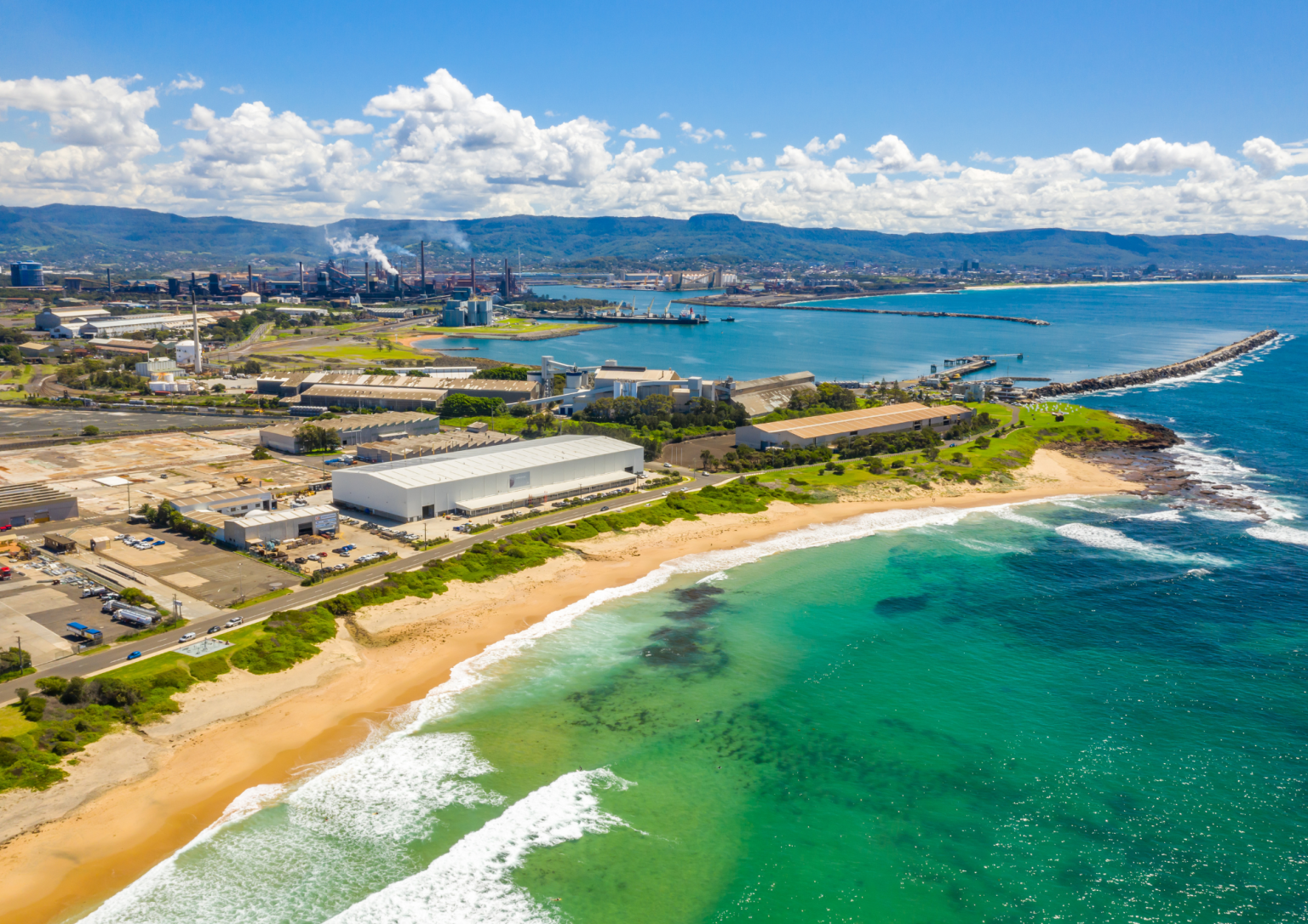Aerial view of a coastal industrial area with factories, warehouses, and a sandy beach leading into vibrant green ocean waters under a partly cloudy sky.
