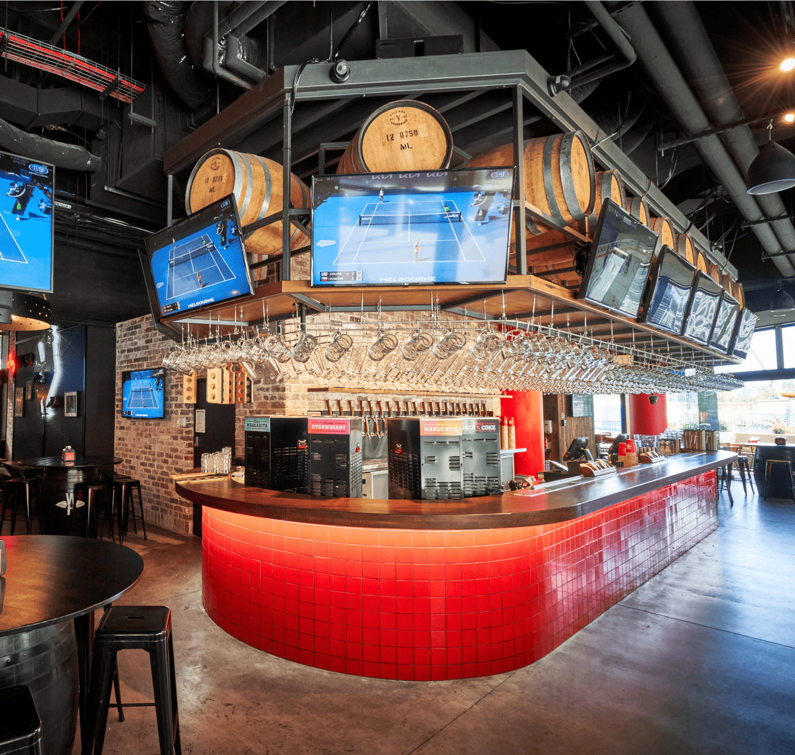 Modern bar interior with red tiled counter, overhead barrels, multiple screens showing a tennis match, and industrial design elements.